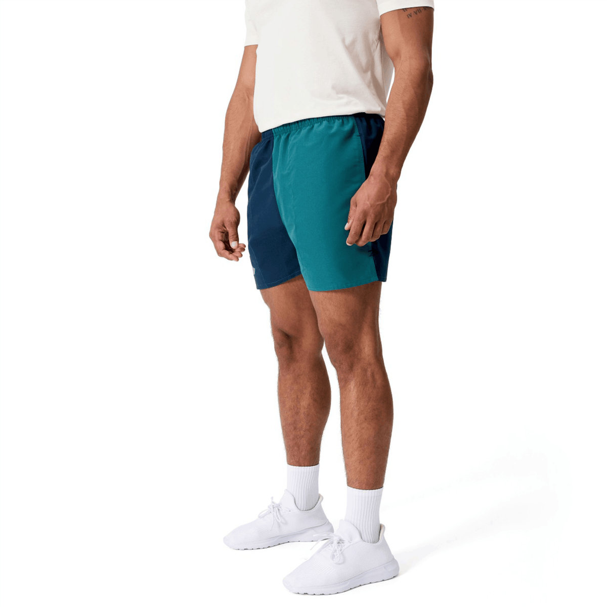 A man wearing a white t-shirt, Canterbury Fundamental Harletic 3″ Rugby Shorts by Canterbury in teal and navy, white socks, and white sneakers, standing on a white background.