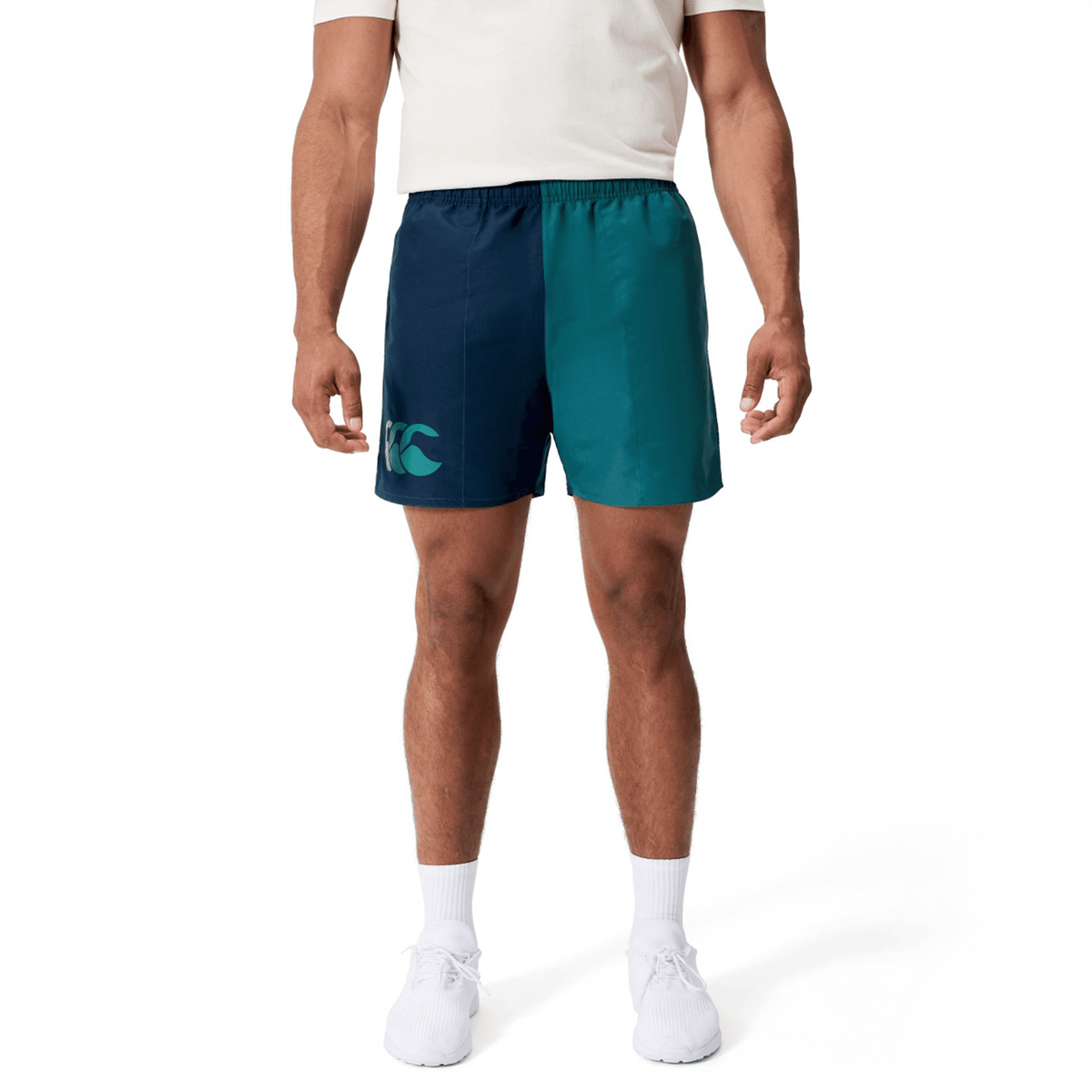 A person wearing a white shirt, white socks, and white sneakers stands against a plain background, paired with Canterbury Fundamental Harletic 3″ Rugby Shorts by Canterbury featuring two-tone blue colors and a logo.
