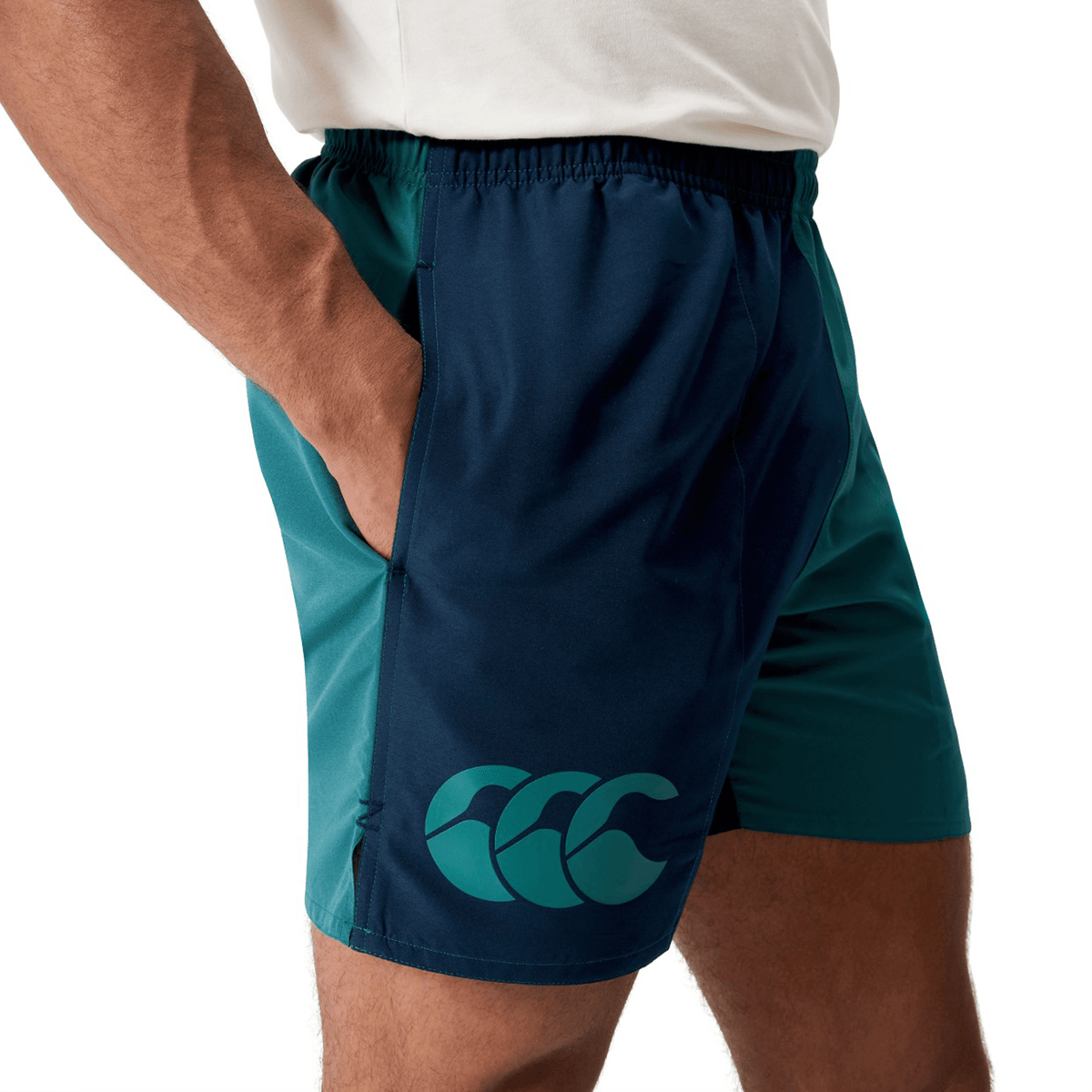 A person wears Canterbury Fundamental Harletic 3″ Rugby Shorts in teal and navy, featuring a wave logo, along with a white t-shirt, standing with one hand in their pocket against a white background.