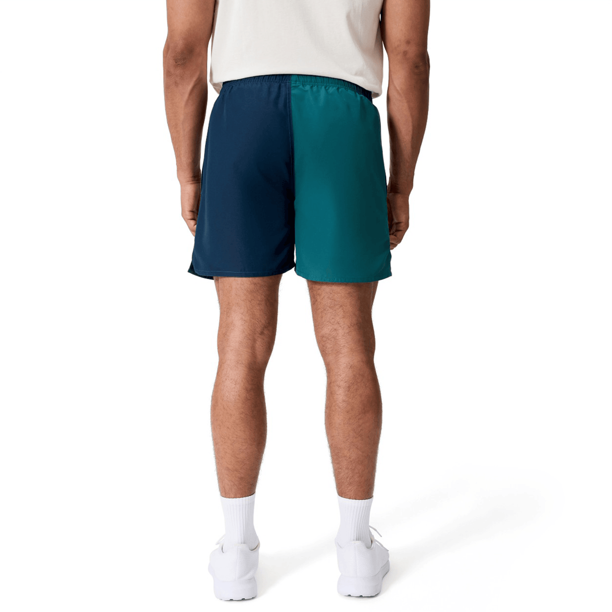 A person stands with their back to the camera, wearing Canterbury Fundamental Harletic 3″ Rugby Shorts in dark blue and teal, paired with a white shirt, white socks, and white sneakers against a plain white background.