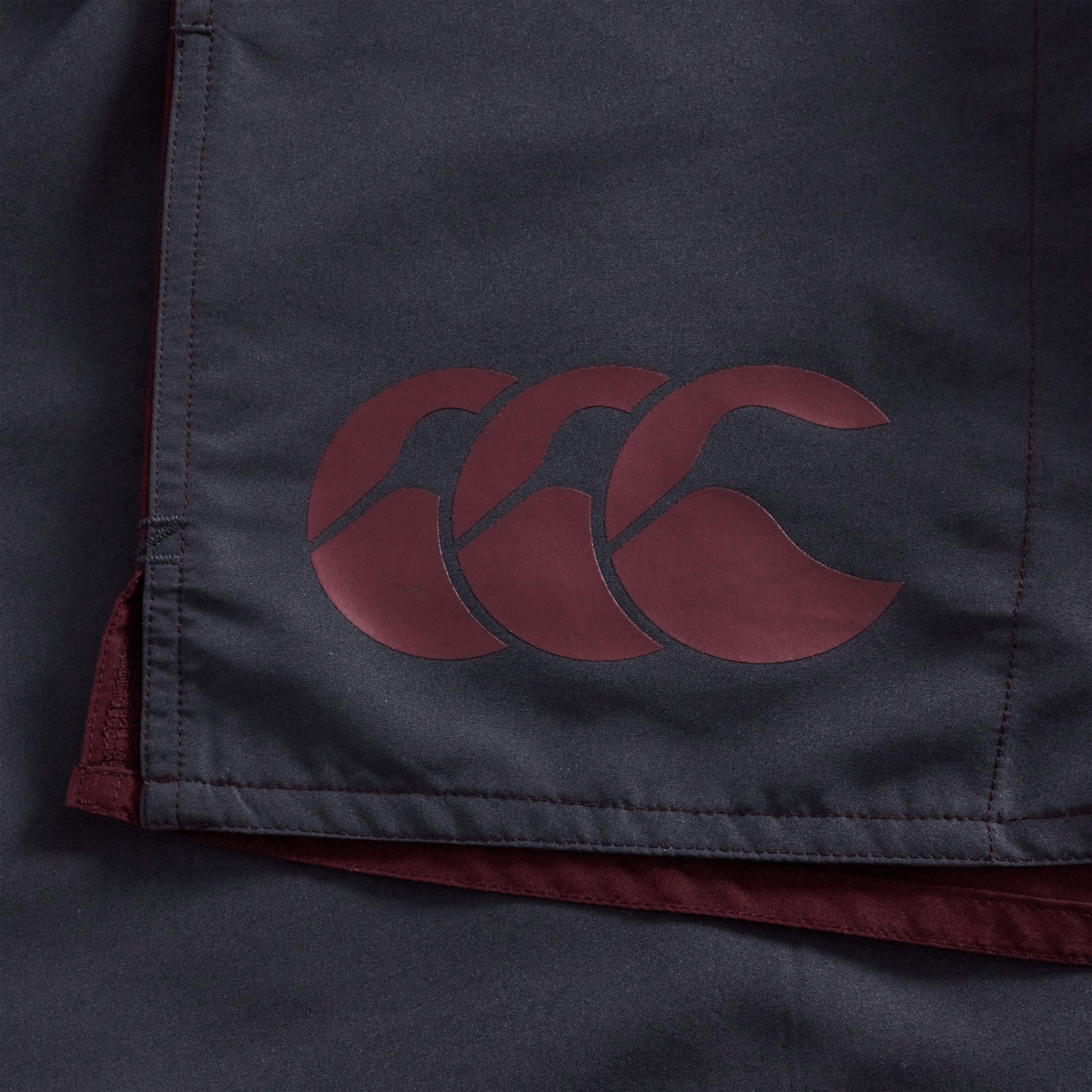 Close-up of a dark fabric featuring a maroon Canterbury logo printed on it, highlighting the garment’s seam and corner detailing—signature elements of the Canterbury Fundamental Harletic 3″ Rugby Shorts by Canterbury.