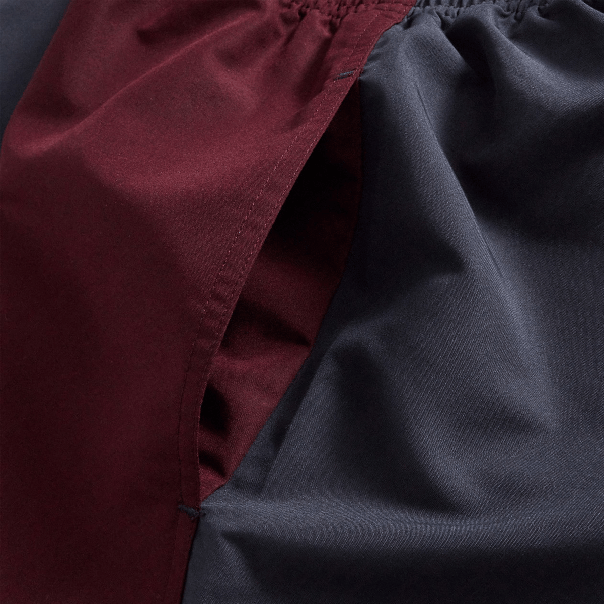 Close-up of fabric with a seam, featuring maroon material on the left and dark gray material on the right—characteristic of the Canterbury Fundamental Harletic 3″ Rugby Shorts by Canterbury—with gathered stitching at the top, a detail commonly found in classic rugby shorts.