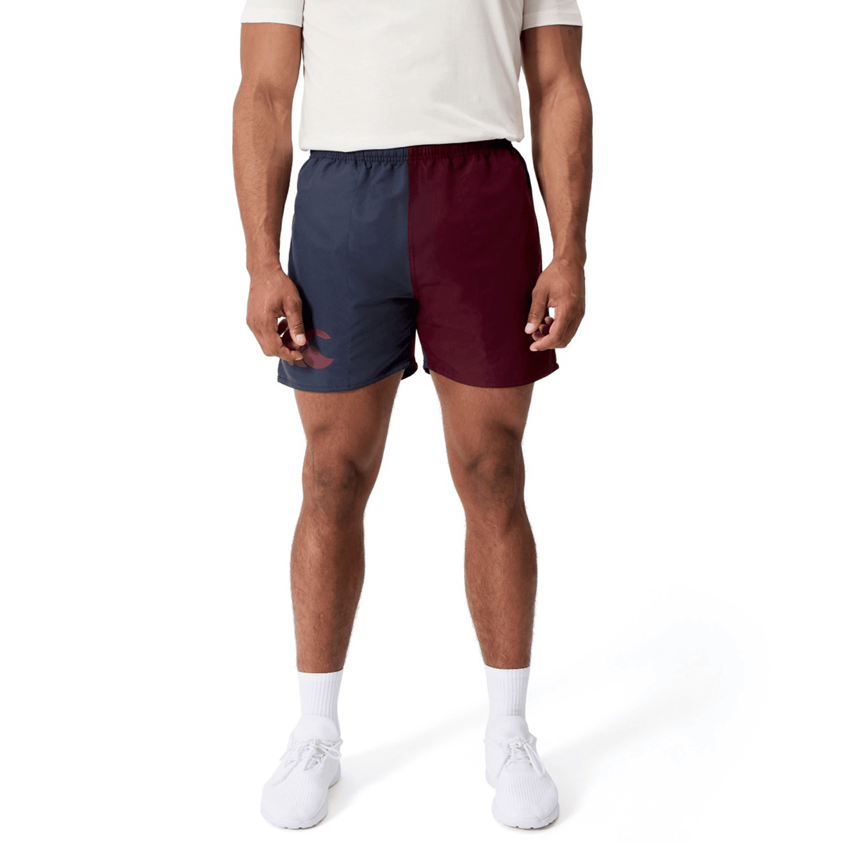 A person stands against a plain white background wearing a white shirt, Canterbury Fundamental Harletic 3″ Rugby Shorts by Canterbury in a color-blocked navy and burgundy design, and white sneakers.