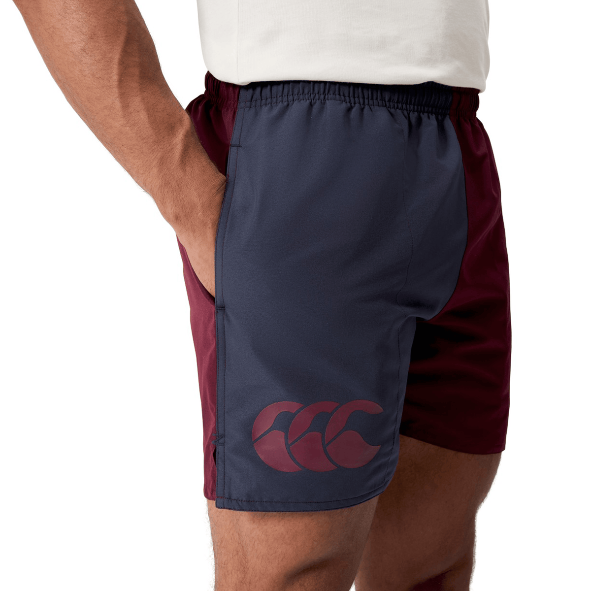 A person is wearing the Canterbury Fundamental Harletic 3″ Rugby Shorts by Canterbury, featuring maroon and navy two-tone panels, a large maroon logo on the left thigh, and a comfortable 3-inch inseam.