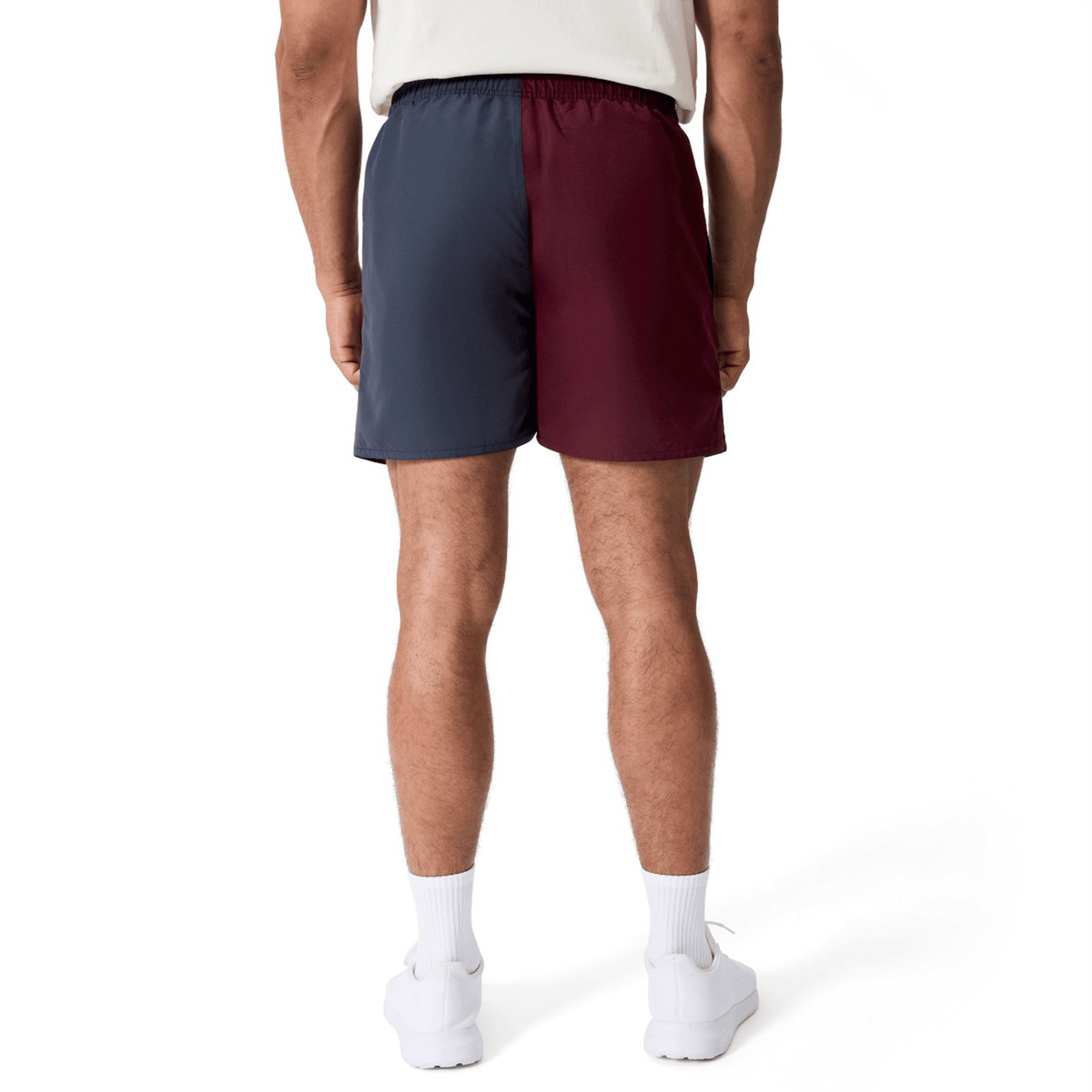 A man stands facing away against a white background, wearing Canterbury Fundamental Harletic 3″ Rugby Shorts by Canterbury—featuring a two-tone design with one navy blue leg and one burgundy leg—paired with a white shirt, white socks, and white sneakers.