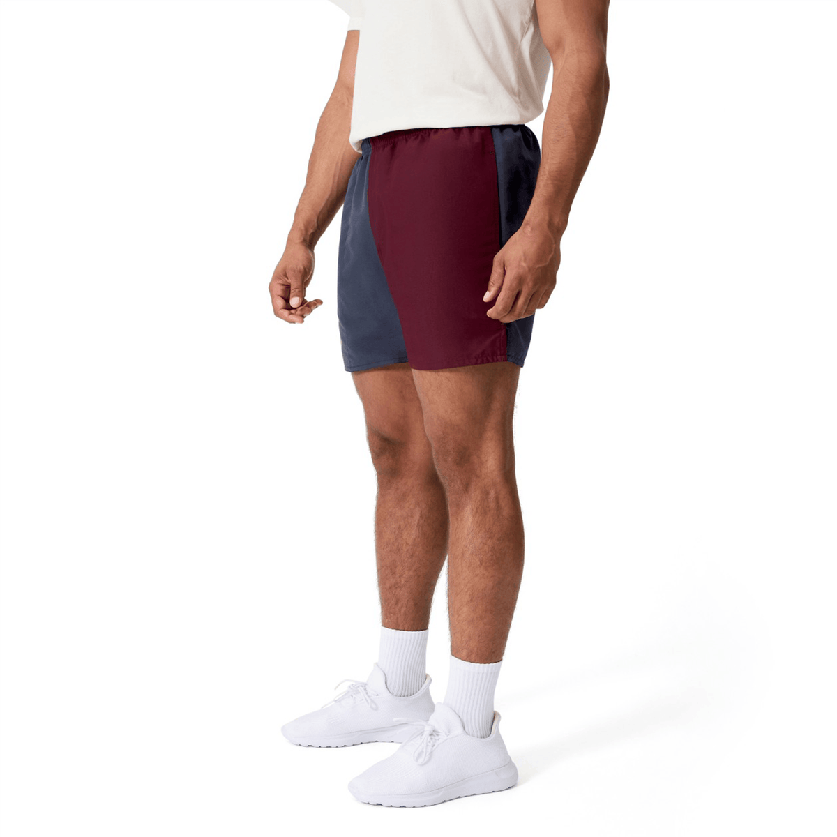 A man wearing a white t-shirt, Canterbury Fundamental Harletic 3″ Rugby Shorts by Canterbury in a color-blocked maroon and navy design, along with white socks and white sneakers, stands against a plain white background.