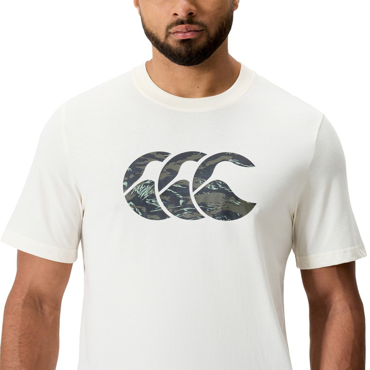 A person wears the Canterbury Force CCC T-Shirt, an athletic cotton-blend white tee with a bold green and brown camo Canterbury CCC logo on the chest.