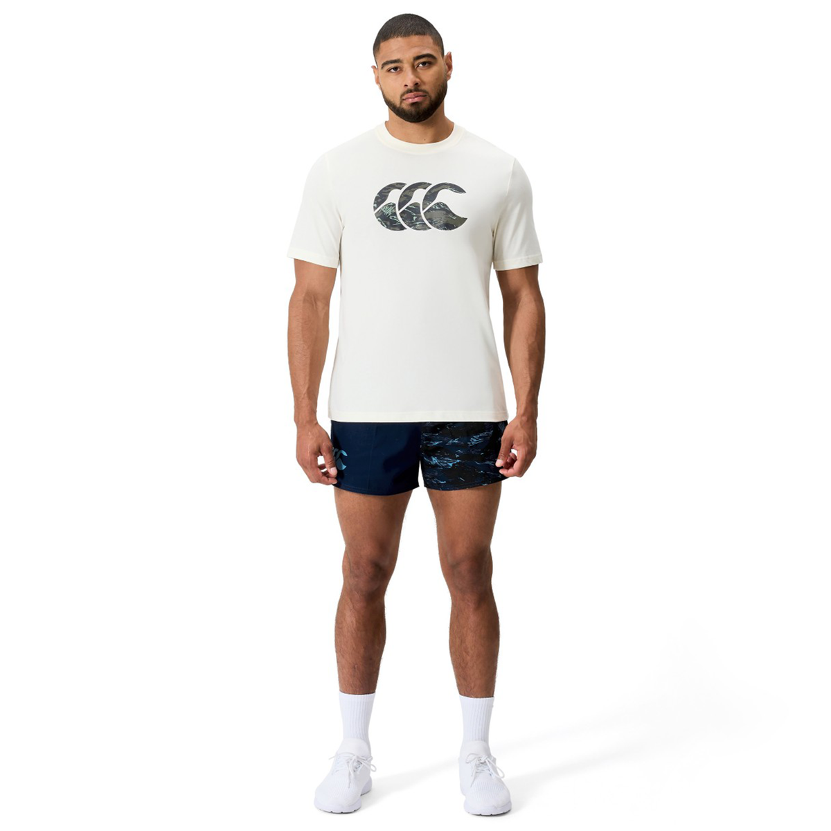 A man faces forward wearing a Canterbury Force CCC T-Shirt, navy patterned shorts, white socks, and white sneakers.