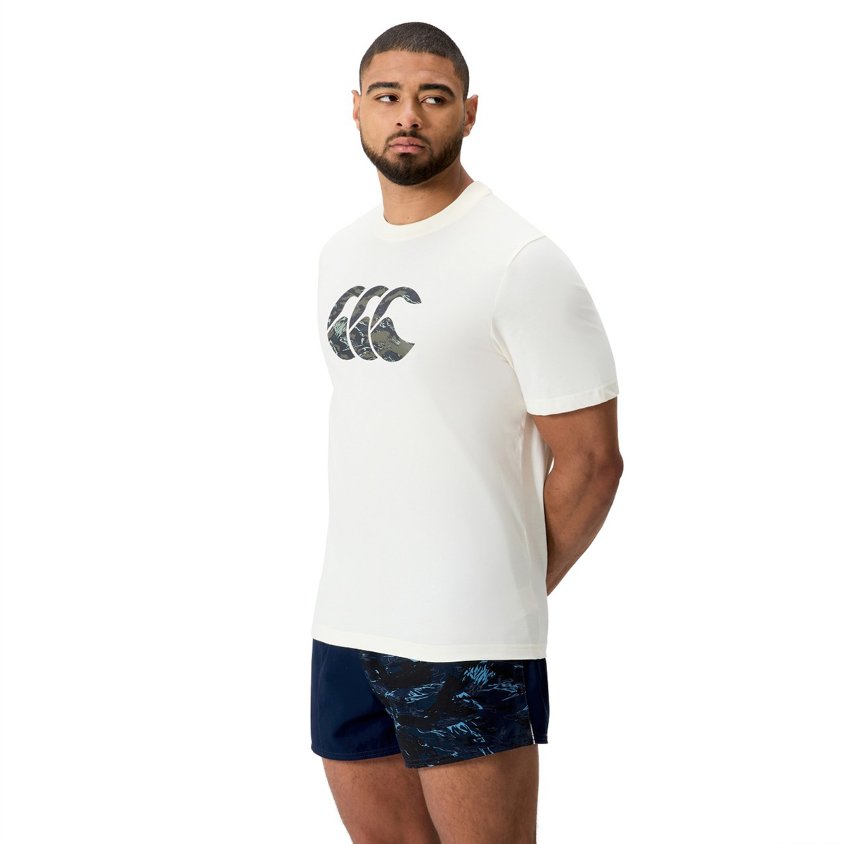 A man in a Canterbury Force CCC T-Shirt with a wave logo and dark patterned shorts stands with his hands behind his back, looking to the side.