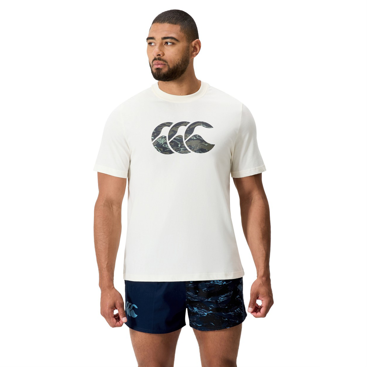 A man wears the Canterbury Force CCC T-Shirt in white with a bold logo and navy cotton-blend shorts, standing forward with his head turned to the side.