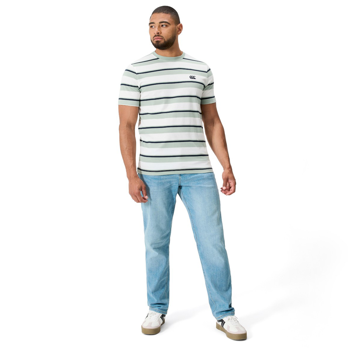 A man wears the Fundamental Yarn Dye Stripe T-Shirt by Canterbury, which features rugby-inspired white and green stripes, styled with light blue jeans and white sneakers against a plain white background.
