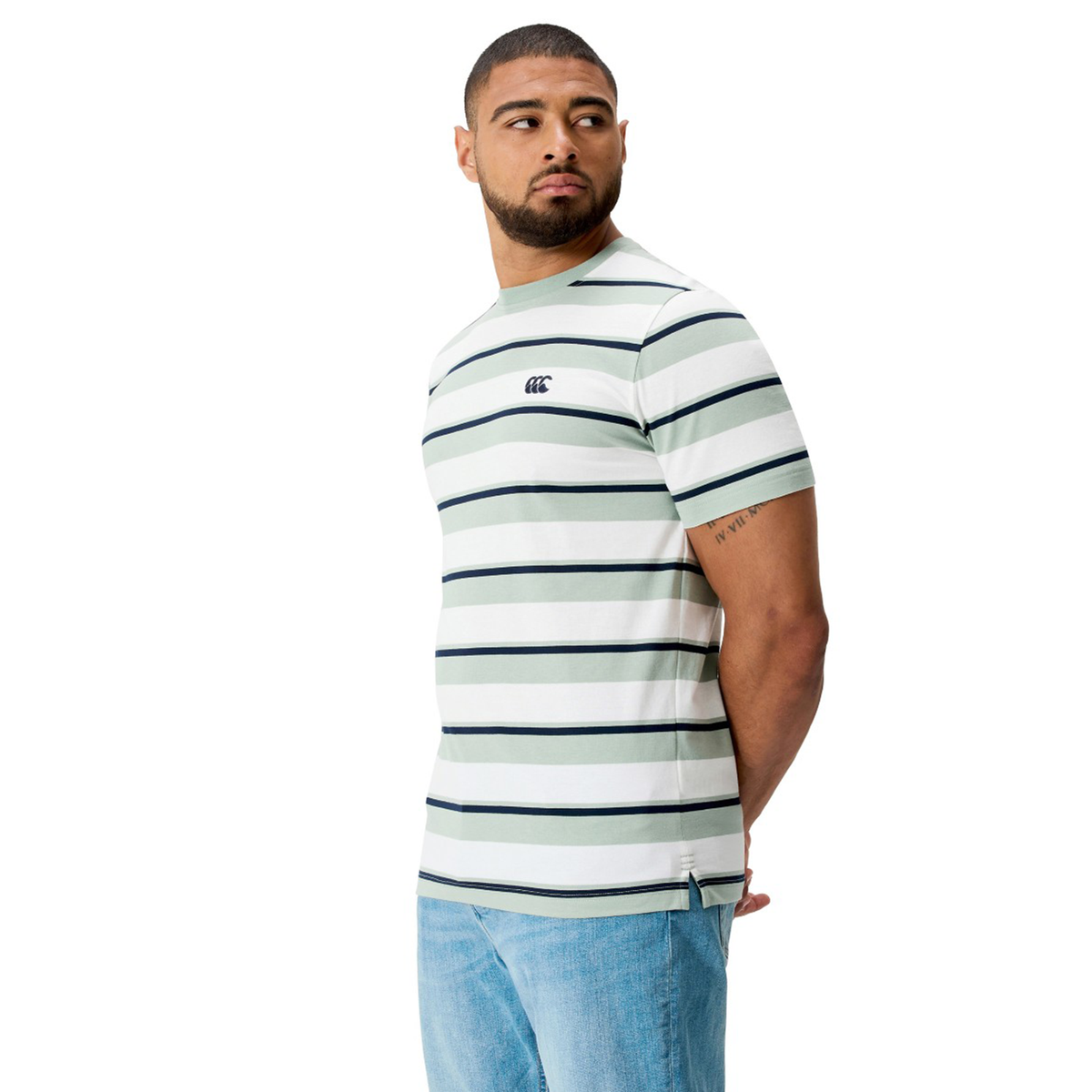 A man in a Canterbury Fundamental Yarn Dye Stripe T-Shirt by Canterbury, featuring navy blue and green rugby-style stripes, and light blue jeans stands facing left against a white background.