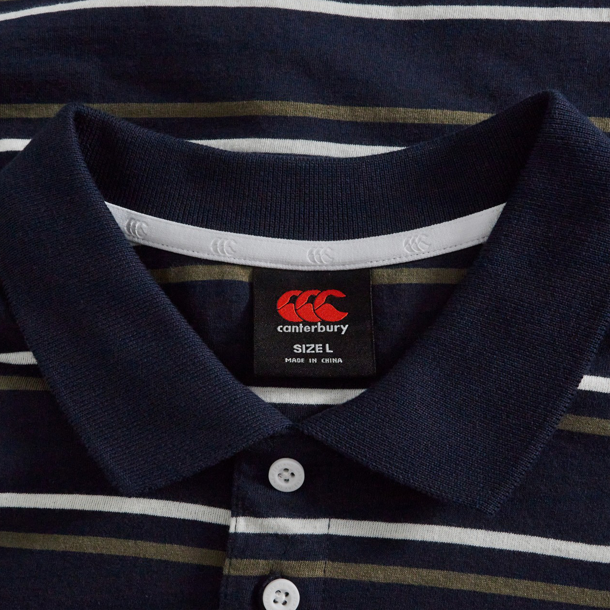 A close-up of the Fundamental Yarn Dye Stripe Polo by Canterbury in navy, showing white buttons and a brand label with size L and "Made in China.