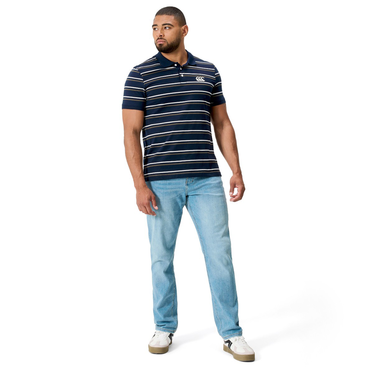 A man wears the Fundamental Yarn Dye Stripe Polo by Canterbury in navy and white, paired with light blue jeans and white sneakers, standing against a white background.