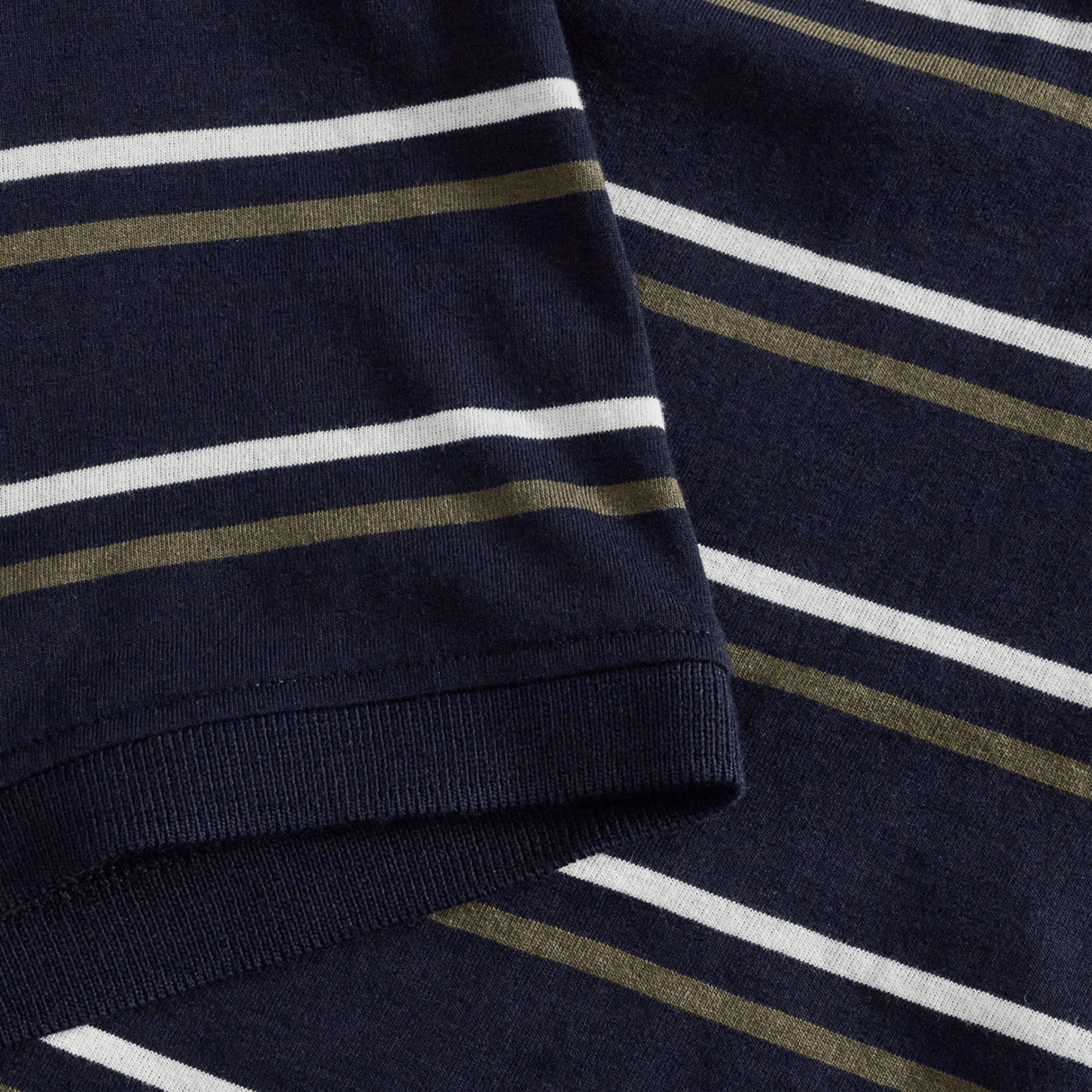 Close-up of a navy blue fabric with horizontal white and olive green stripes, characteristic of the Canterbury Yarn Dye Stripe Polo. The image features a sleeve overlapping the main body of the garment, evoking classic Canterbury rugby polo styling.