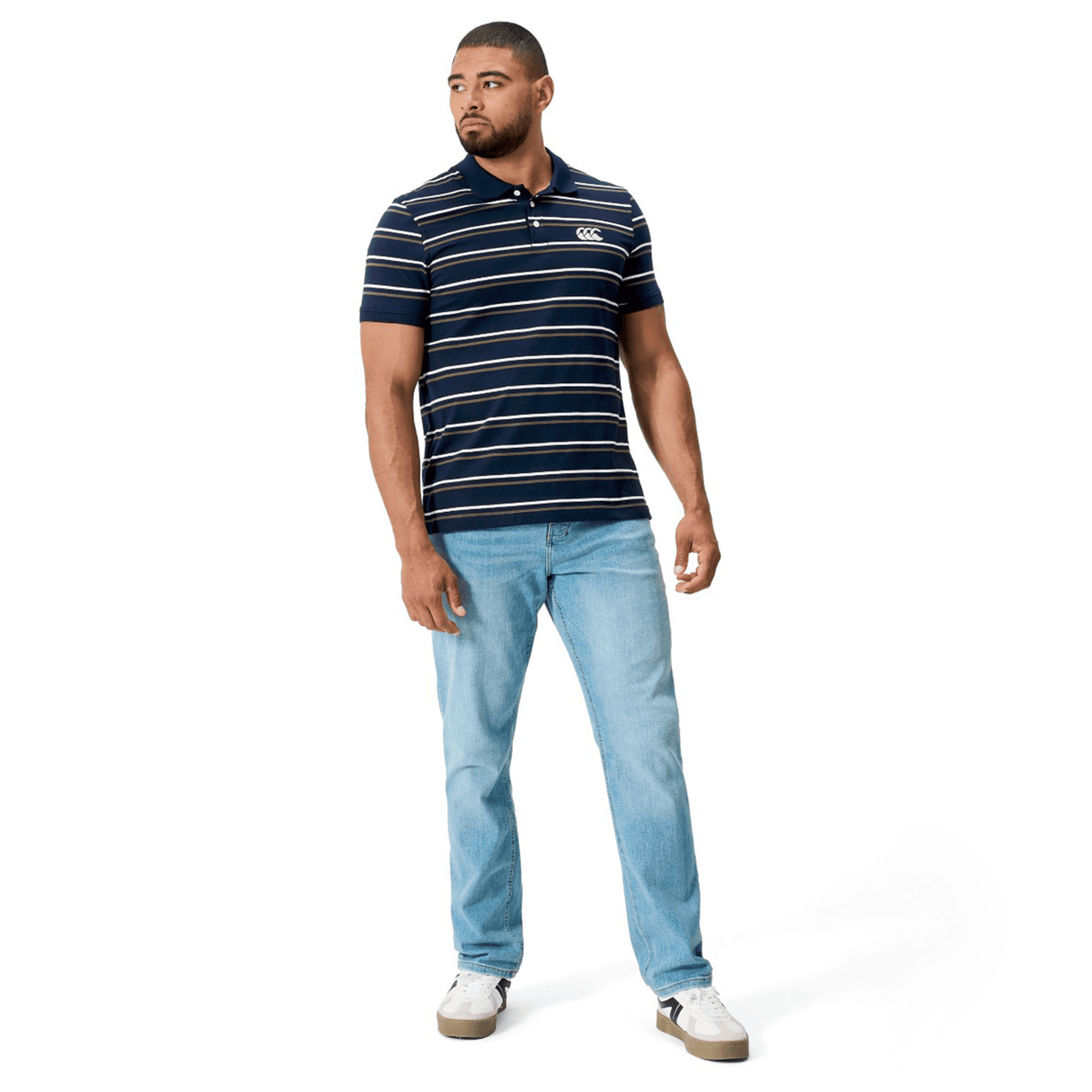 A man stands against a white background, wearing the Canterbury Yarn Dye Stripe Polo by Canterbury, paired with light blue jeans and white sneakers. He looks to his left with his arms relaxed at his sides.