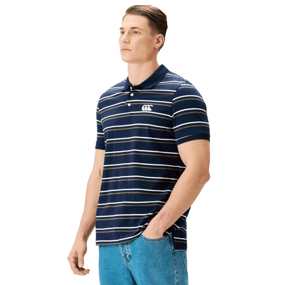 A man wearing the Canterbury Yarn Dye Stripe Polo by Canterbury, featuring navy blue and white stripes and a logo on the chest, paired with blue jeans and standing with one hand in his pocket.