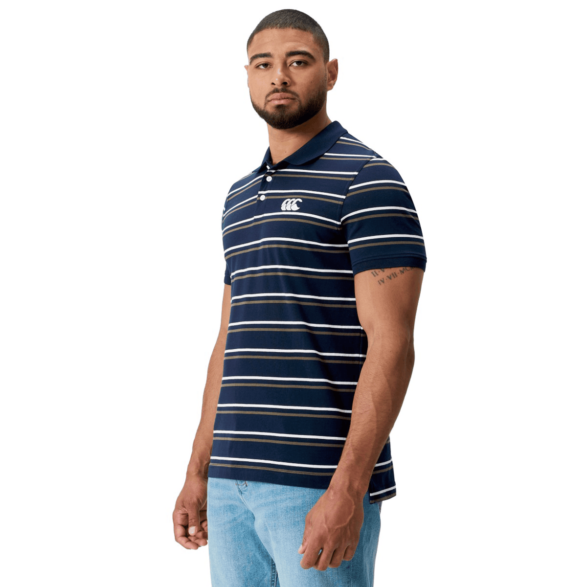 A man is wearing a Canterbury Yarn Dye Stripe Polo by Canterbury in navy blue and white with short sleeves, paired with blue jeans, standing against a plain white background.
