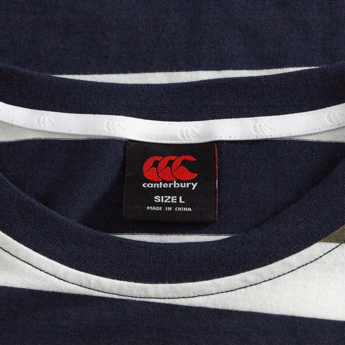 Close-up of the Fundamental Yarn Dye Stripe T-Shirt by Canterbury in navy blue and white, rugby-inspired, highlighting the neck area and black label showing size L and "Made in China" text.