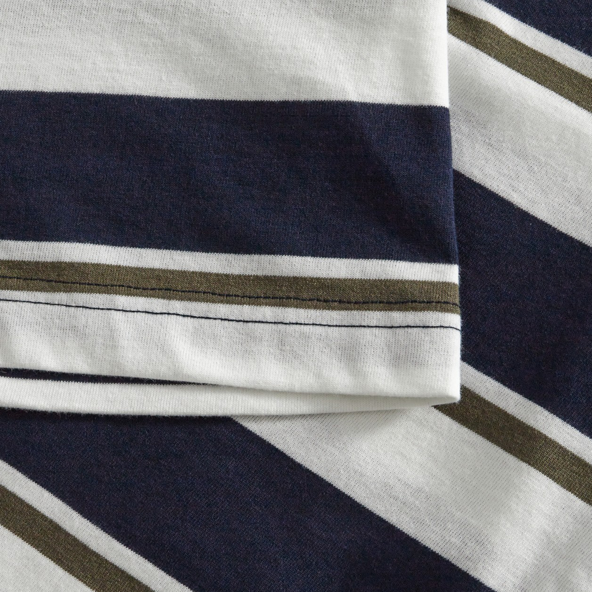 Close-up of Canterbury’s Fundamental Yarn Dye Stripe T-Shirt fabric shows thick navy and white diagonal stripes with a thin olive stripe, plus stitched hems, reflecting its rugby-inspired style.