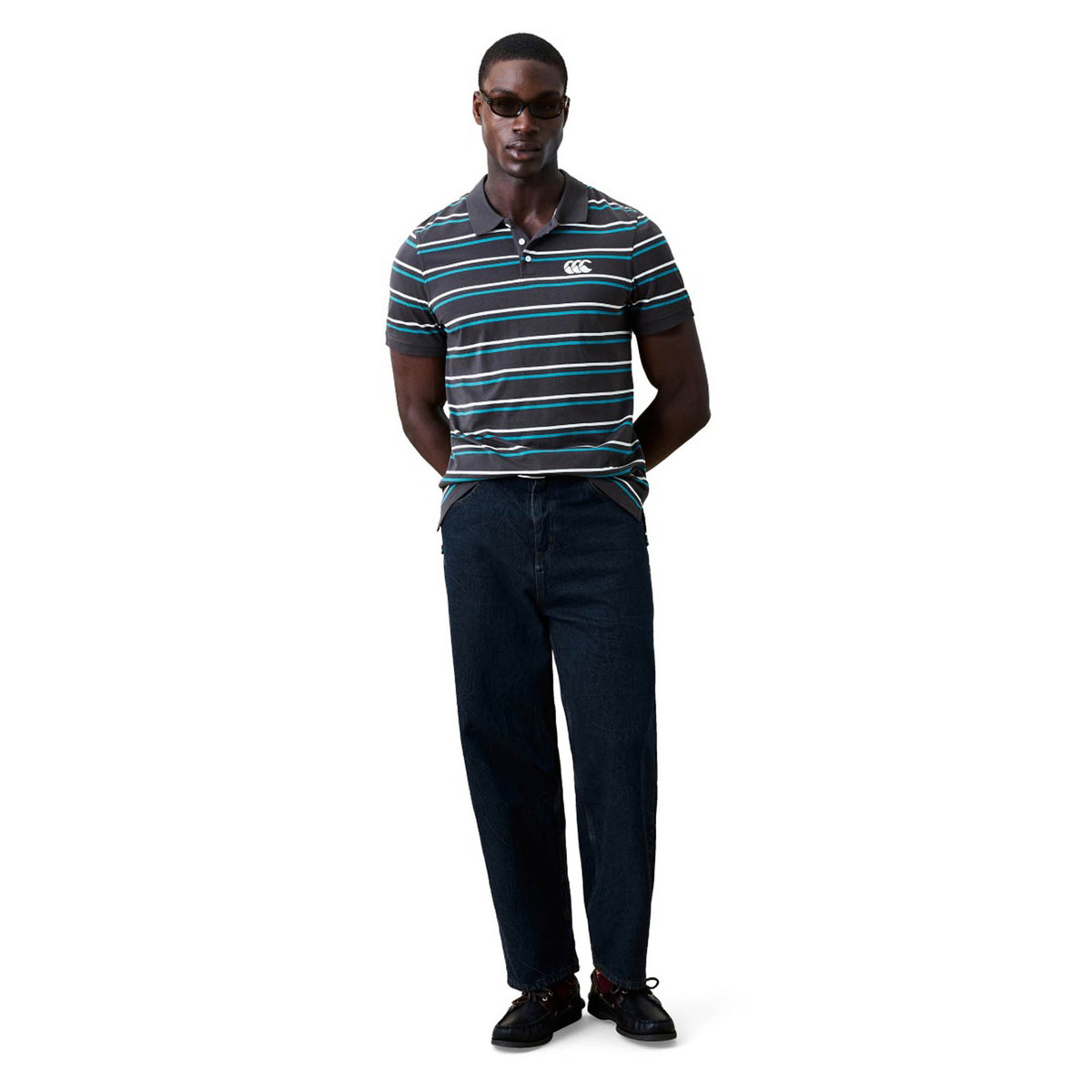 A man wearing sunglasses, a Canterbury Yarn Dye Stripe Polo by Canterbury, dark jeans, and black shoes stands with his hands behind his back against a plain white background.