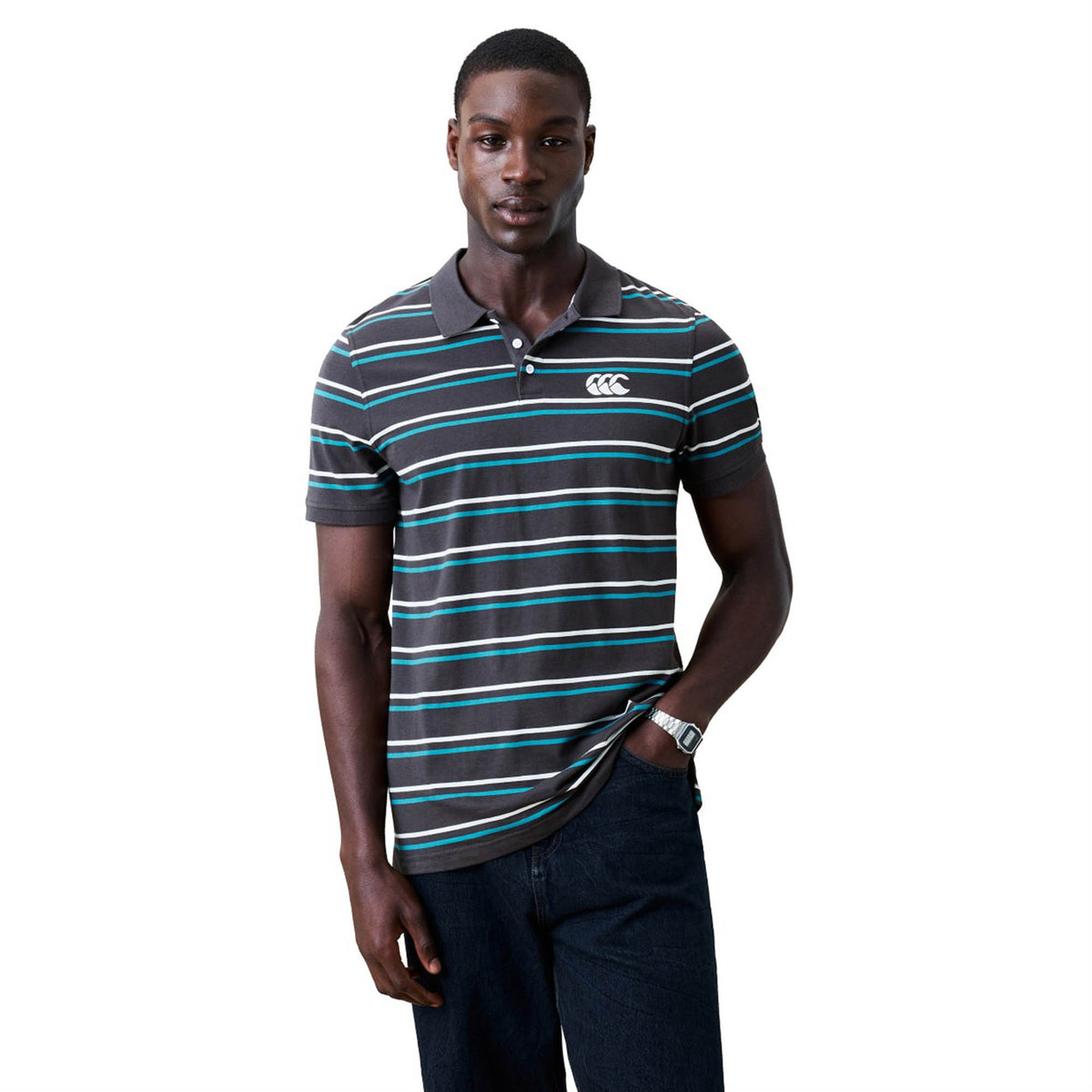 A man stands against a white background wearing the Canterbury Yarn Dye Stripe Polo from Canterbury, inspired by rugby heritage, paired with dark jeans and with one hand in his pocket.