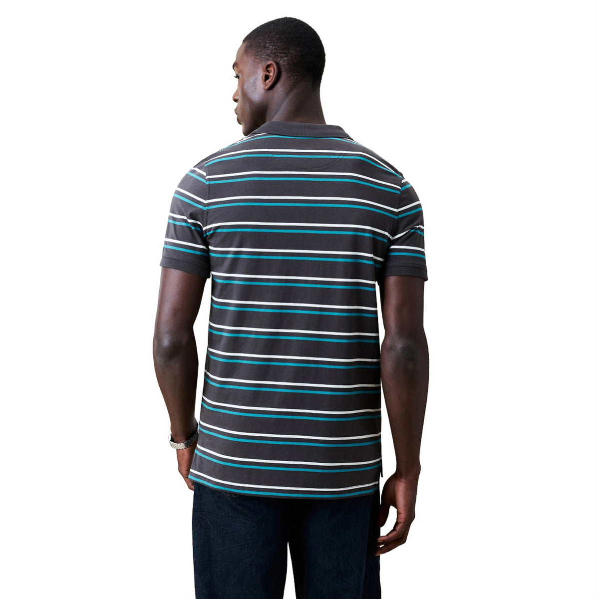 A man stands with his back to the camera, dressed in a Canterbury Yarn Dye Stripe Polo by Canterbury. The short-sleeve polo features horizontal stripes in white, blue, and gray tones and is paired with dark pants, set against a plain white background.