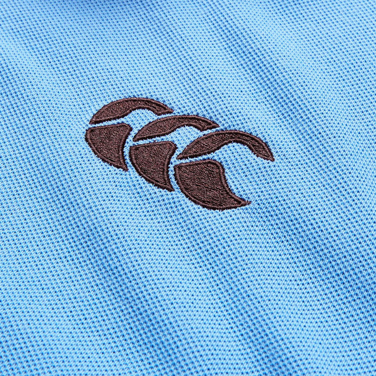 Close-up of a blue textured fabric from the Canterbury Solid Dye Polo, featuring a black embroidered Canterbury logo with three overlapping curved shapes.