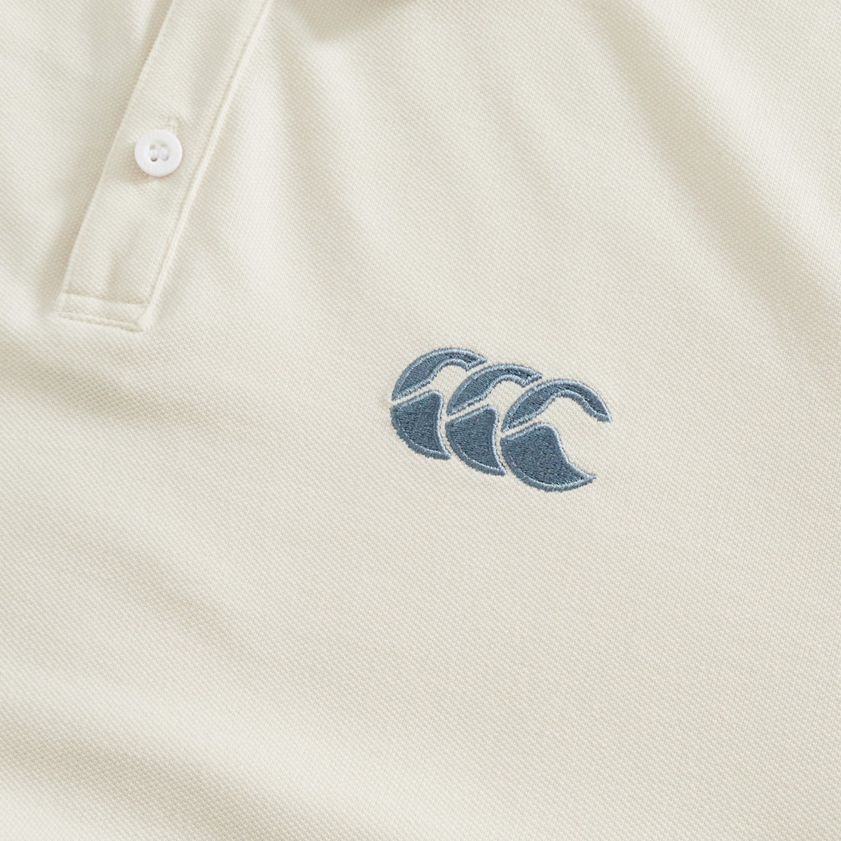 Close-up of the cream-colored Fundamental Solid Dye Polo by Canterbury, featuring a stitched blue logo with three overlapping waves on the chest, showcasing its subtle polo and rugby-inspired design.