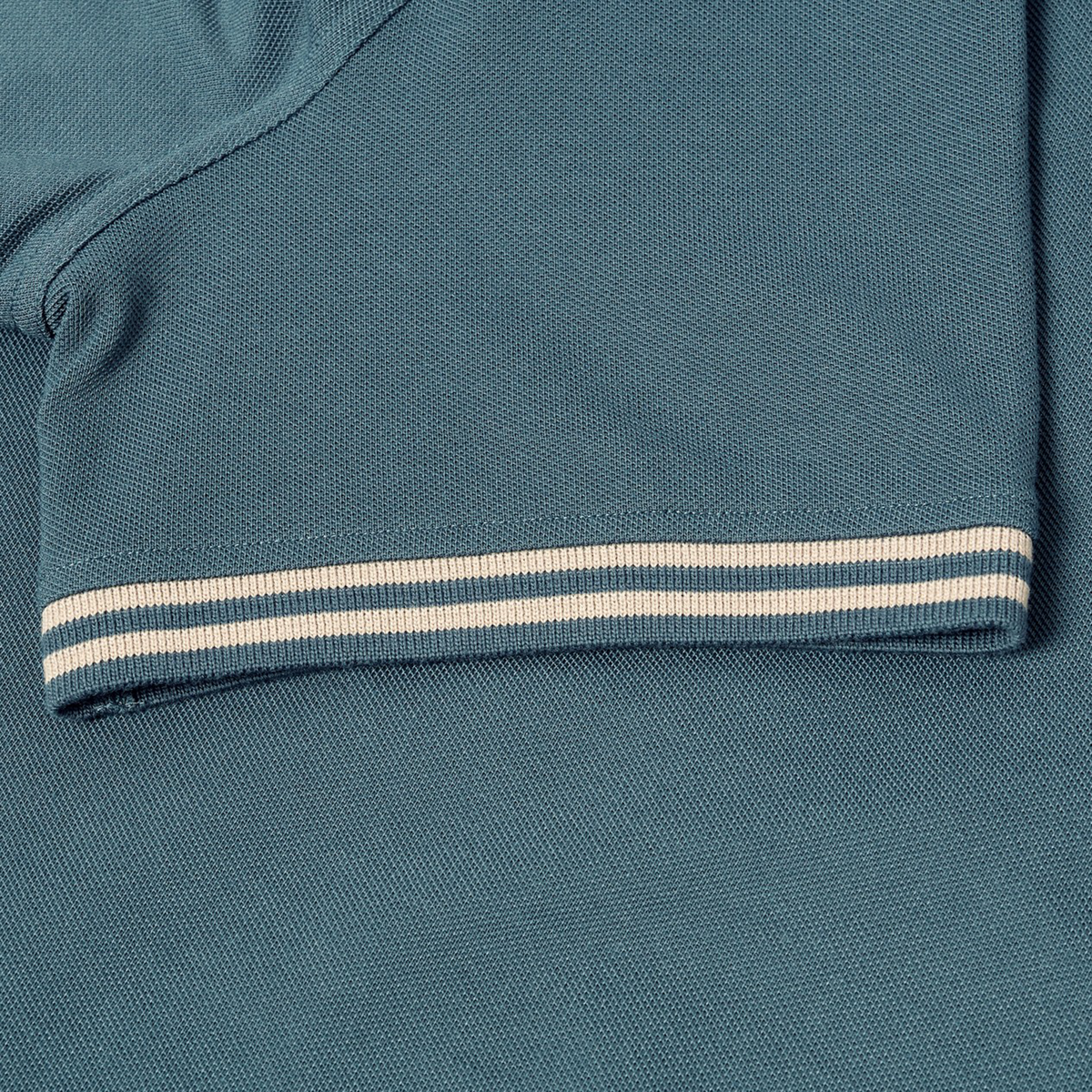 Close-up of the Canterbury Solid Dye Polo by Canterbury, featuring short sleeves in a rich teal color and a beige and white striped cuff, highlighting its rugby-inspired athletic style with detailed fabric texture and precise stitching.