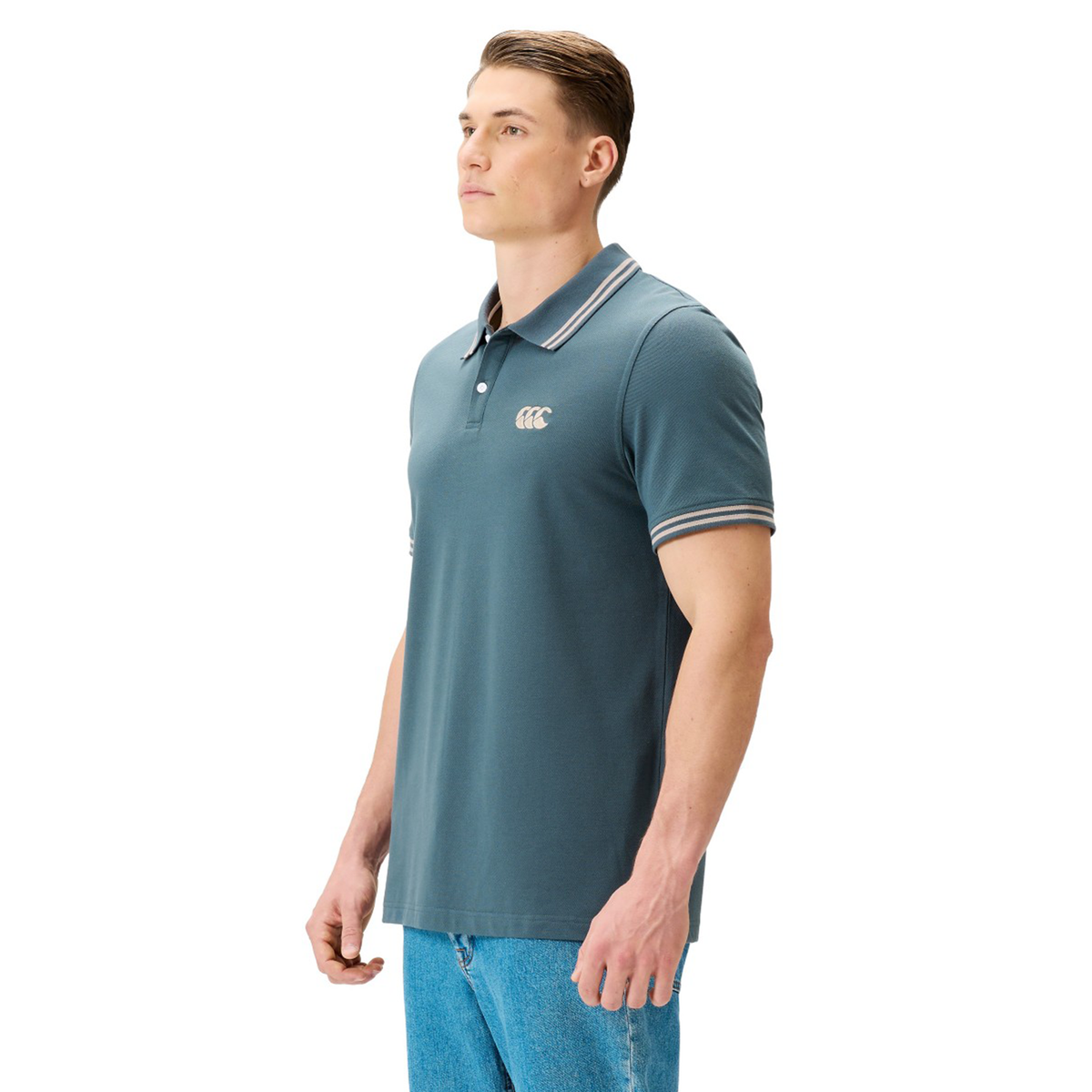 A man stands facing slightly to the side against a plain white background, wearing the Canterbury Solid Dye Polo by Canterbury—a rugby-inspired short-sleeve teal polo shirt with a logo on the chest—paired with blue jeans.