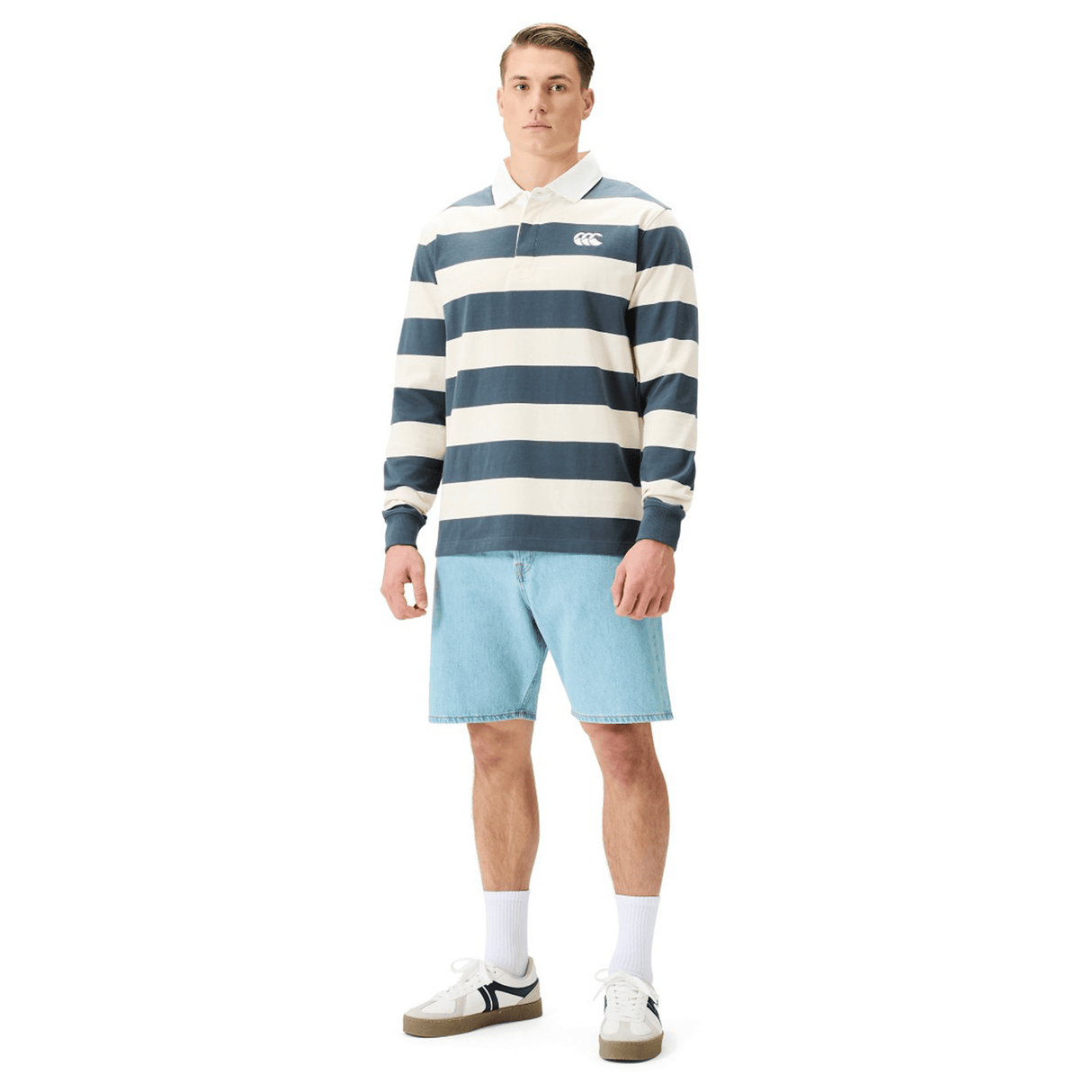 A man wears the Canterbury Yarn Stripe Rugby Jersey by Canterbury, featuring navy and white stripes, paired with light blue denim shorts, white socks, and white sneakers against a plain white background.