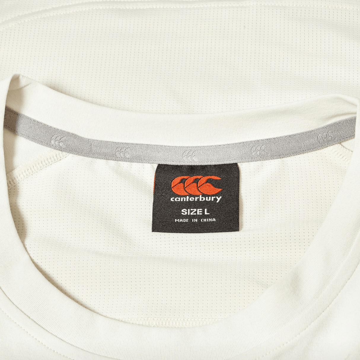Close-up of a white Canterbury Everest Dart Tech T-Shirt featuring a black and orange Canterbury label indicating size L and made in China.