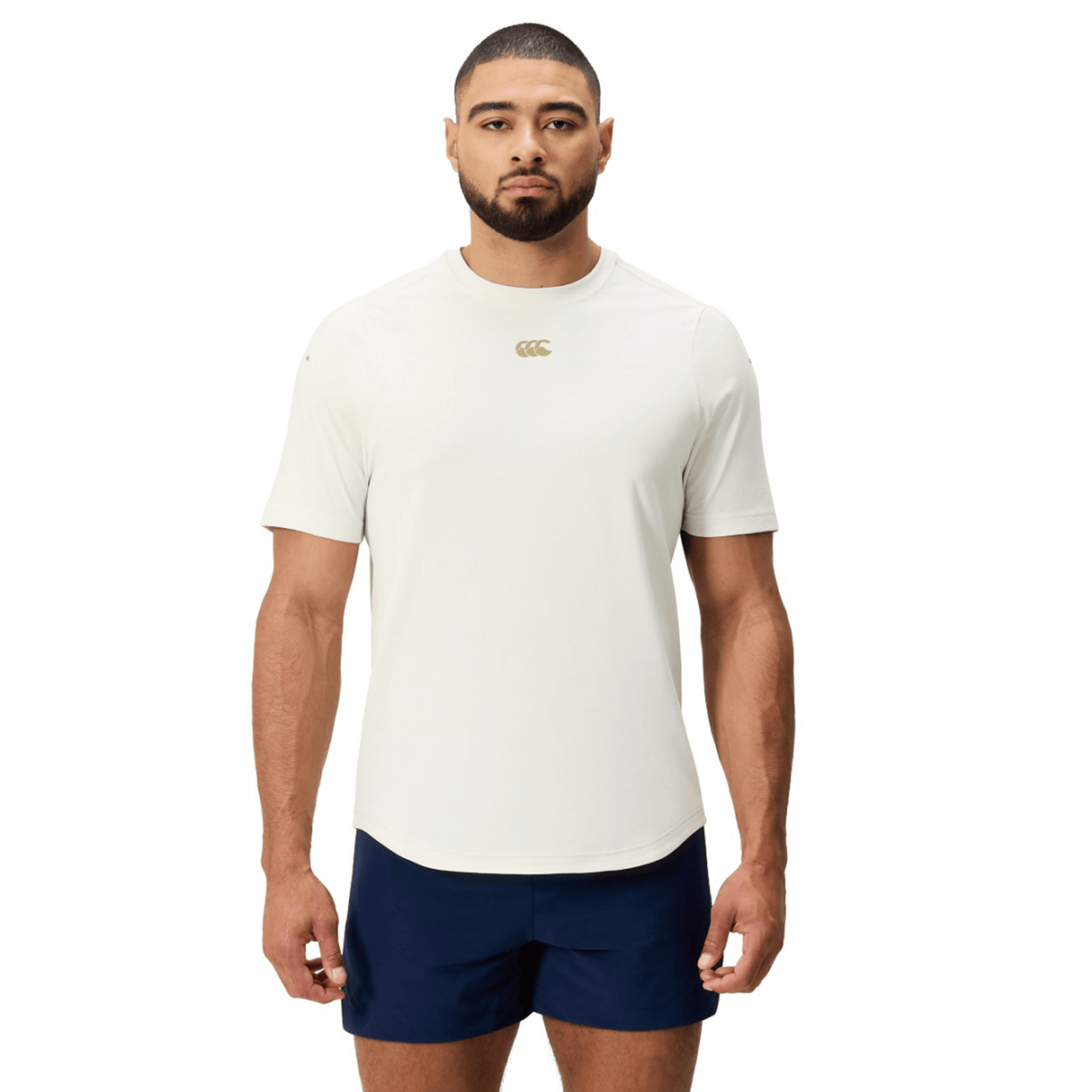 A man stands facing forward, wearing a Canterbury Everest Dart Tech T-Shirt in white with a small gold logo and navy blue shorts, against a plain white background.