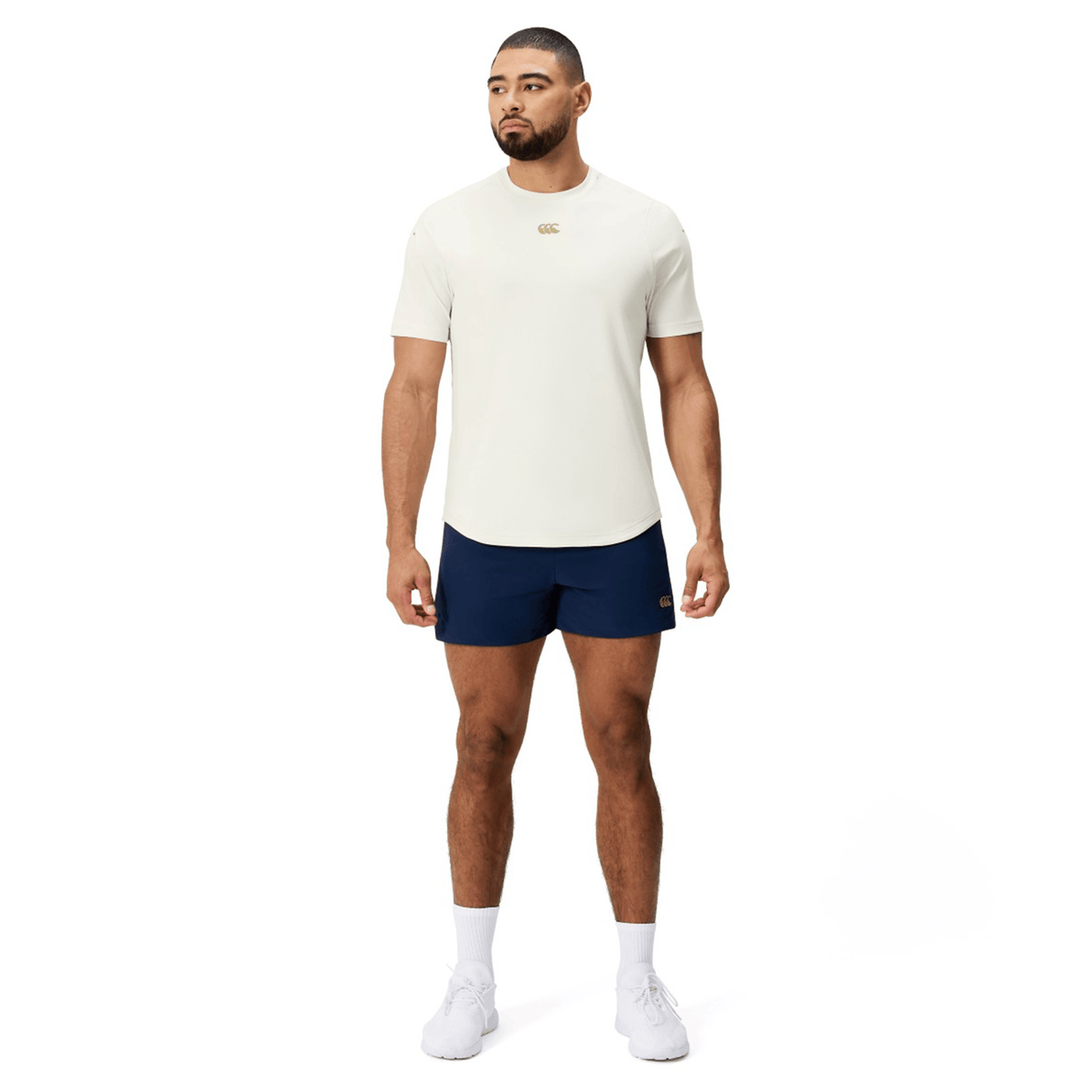 A man faces forward wearing the Canterbury Everest Dart Tech T-Shirt in cream, navy blue shorts, white socks, and white sneakers against a plain white background.