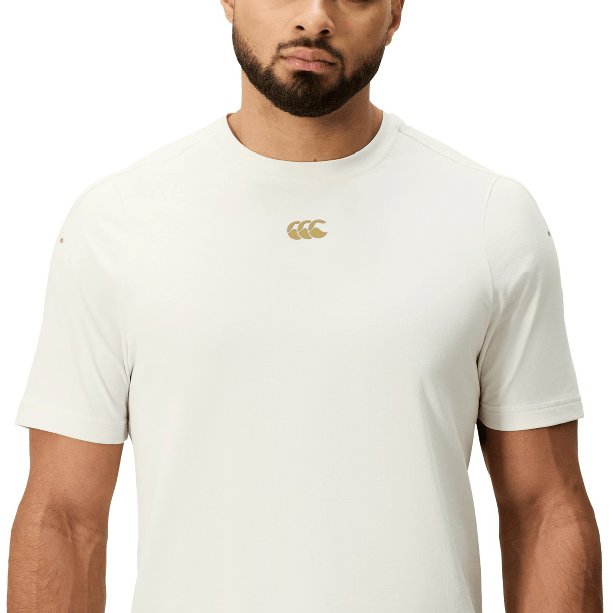 A man wears the Canterbury Everest Dart Tech T-Shirt by Canterbury, a fitted off-white quick-drying short-sleeve top with a small gold chest logo, shown from shoulders to waist against a plain background.