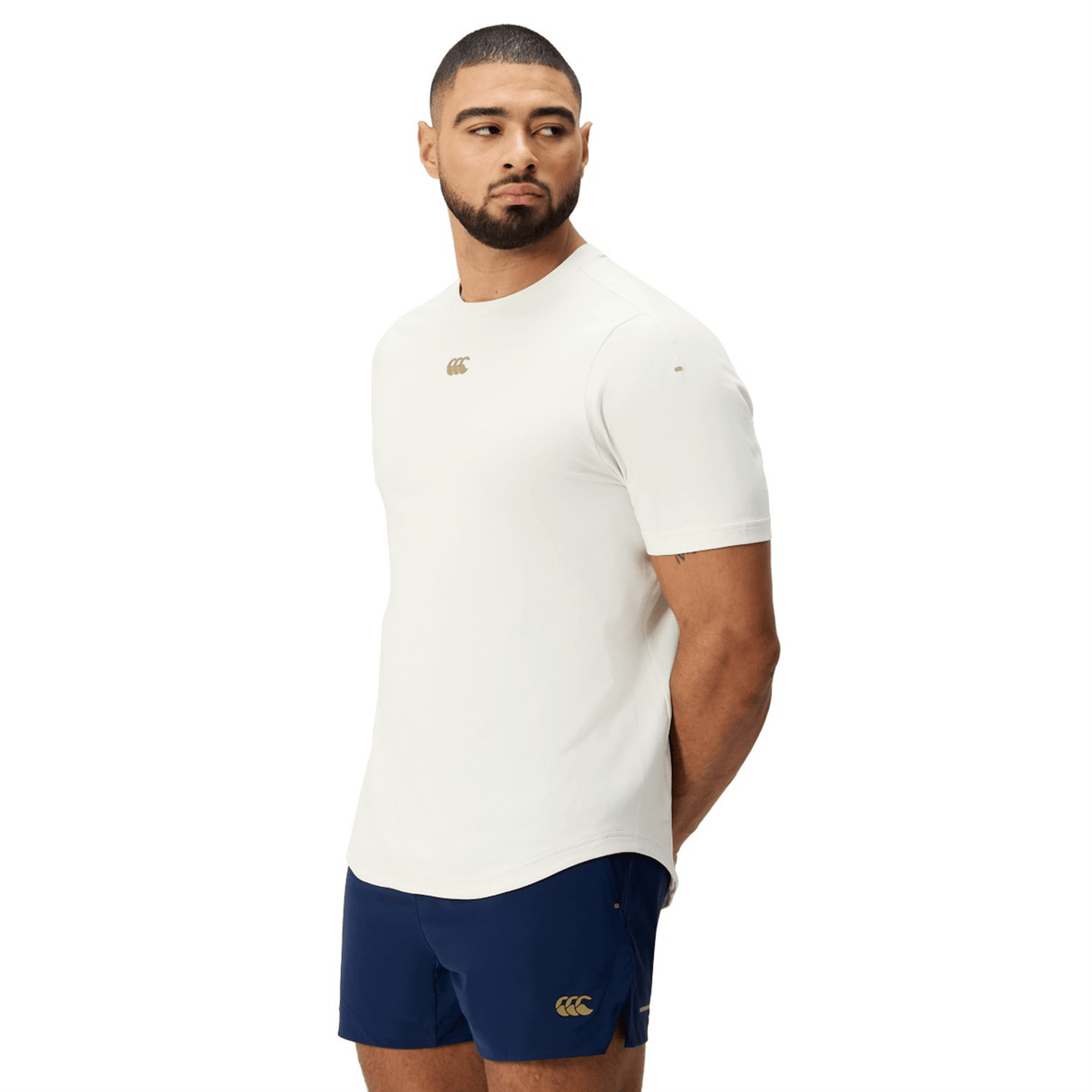 A man stands slightly angled with his hands behind his back, wearing a white Canterbury Everest Dart Tech T-Shirt by Canterbury and navy blue shorts against a plain white background.