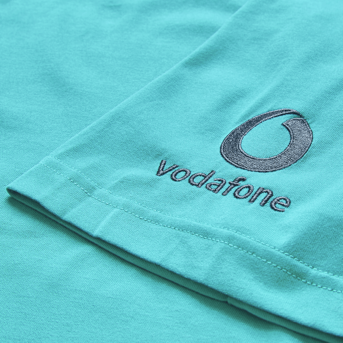 Close-up of the Ireland 26/27 Summer Tour Team Cotton T-Shirt by Canterbury, featuring embroidered Vodafone logo and text on turquoise fabric.