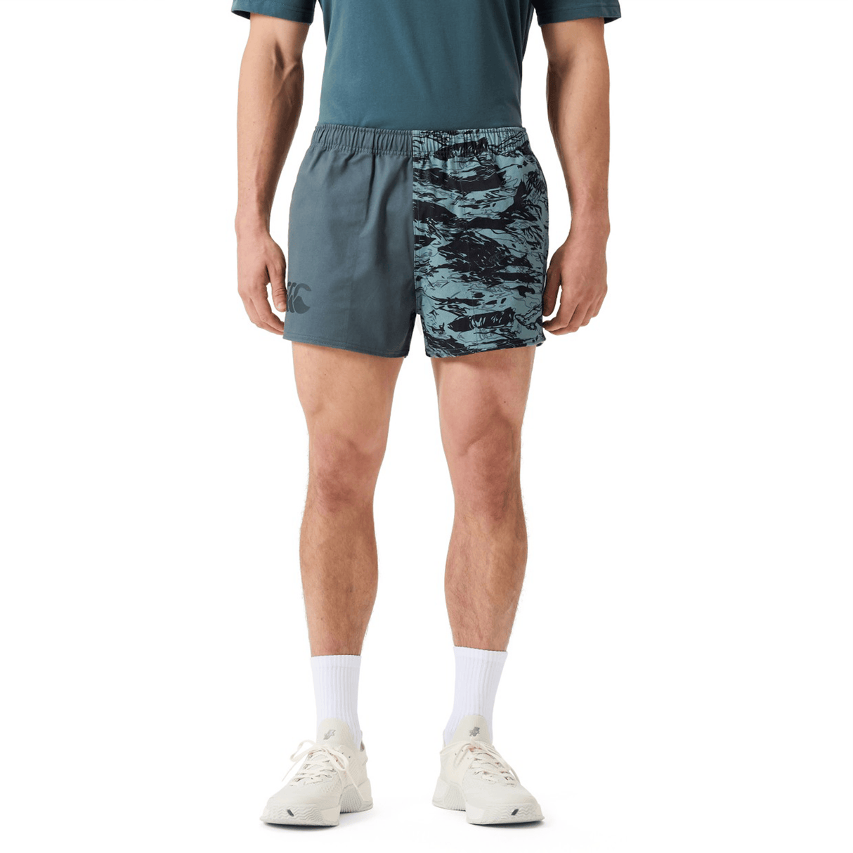 A person wearing Canterbury Force Harlequin Drill Shorts by Canterbury in a teal pattern, a dark green shirt, white socks, and white sneakers stands against a plain white background.