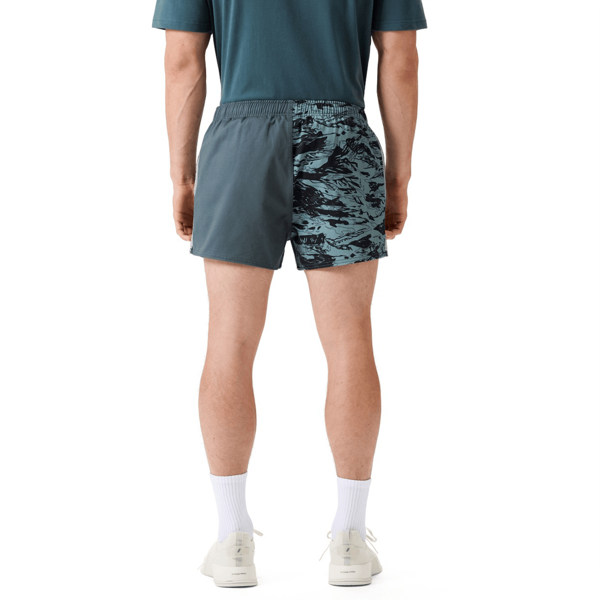 A man stands with his back to the camera, wearing a teal shirt, Canterbury Force Harlequin Drill Short by Canterbury, white socks, and white sneakers.
