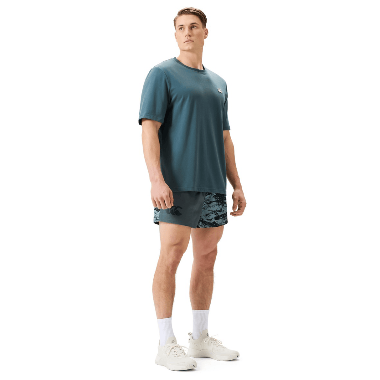 A man stands against a plain white background wearing a teal athletic shirt, Canterbury Force Harlequin Drill Short by Canterbury, white socks, and white sneakers.
