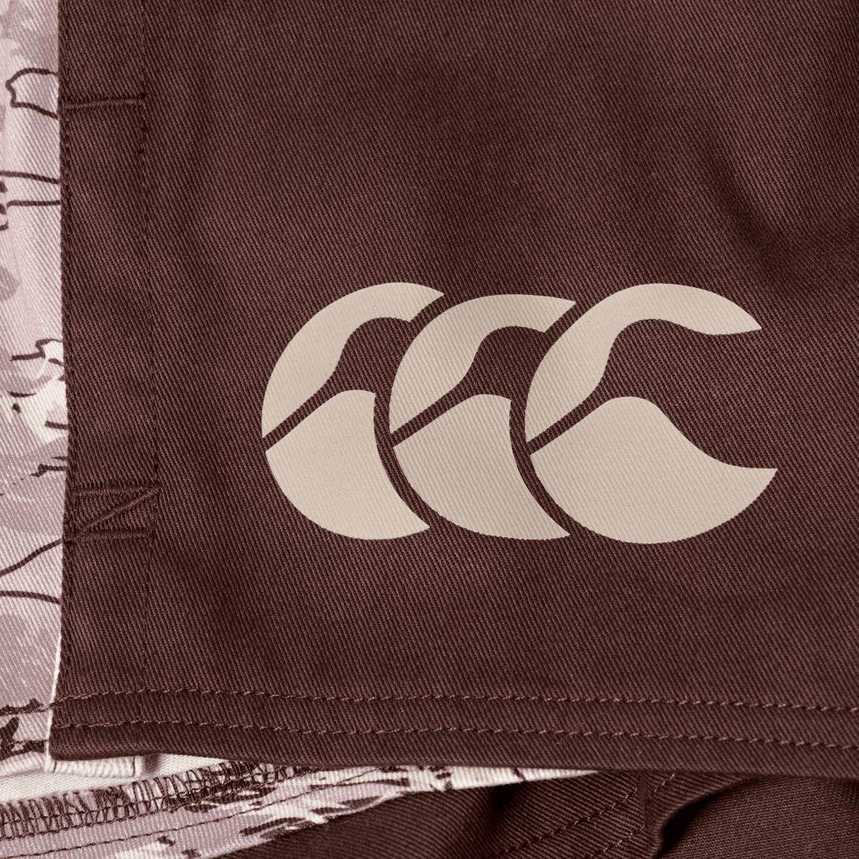 Close-up of brown fabric with a beige triple curved logo, likely from the Canterbury Force Harlequin Drill Short by Canterbury.