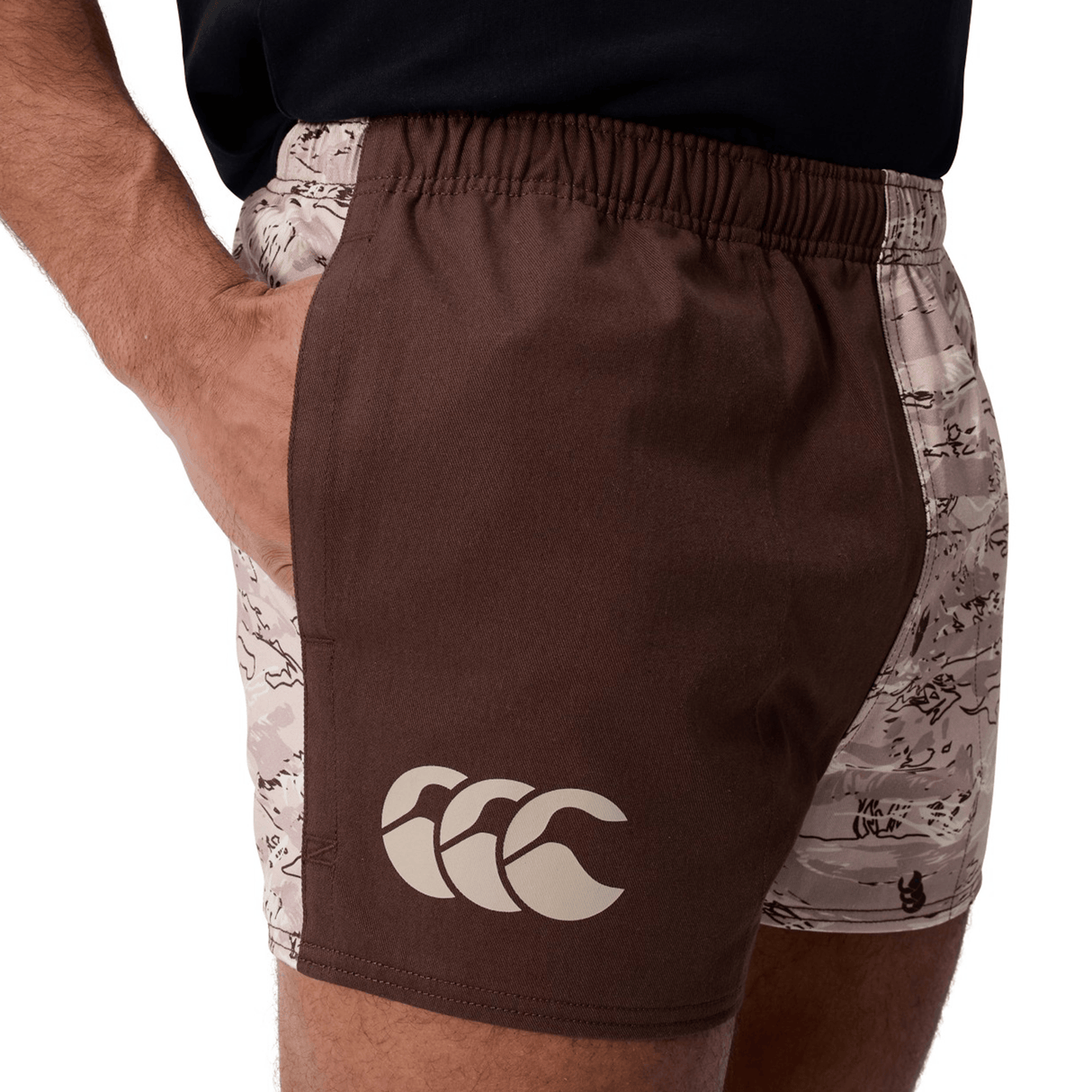 Someone wearing Canterbury Force Harlequin Drill Shorts in brown and beige, with a large white Canterbury CCC logo on the left leg and a hand in the pocket.