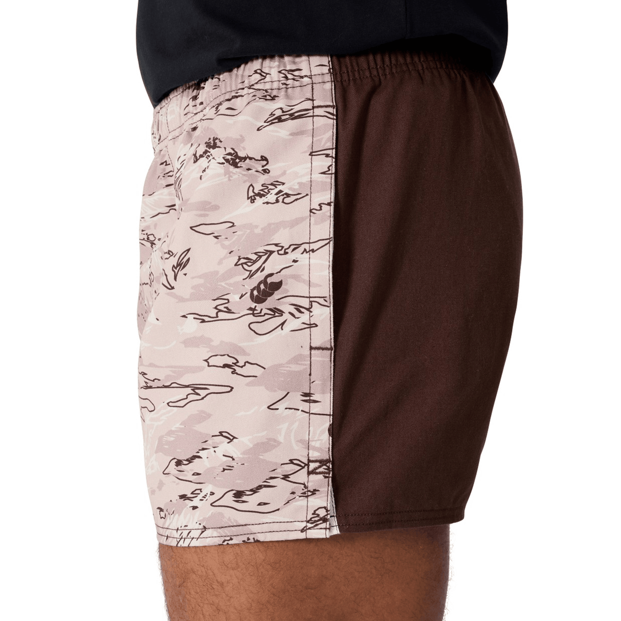 A person wears Canterbury Force Harlequin Drill Shorts by Canterbury, shown from waist to mid-thigh side view; one side features brown camouflage and the other is solid brown, highlighting their military-inspired utility style.