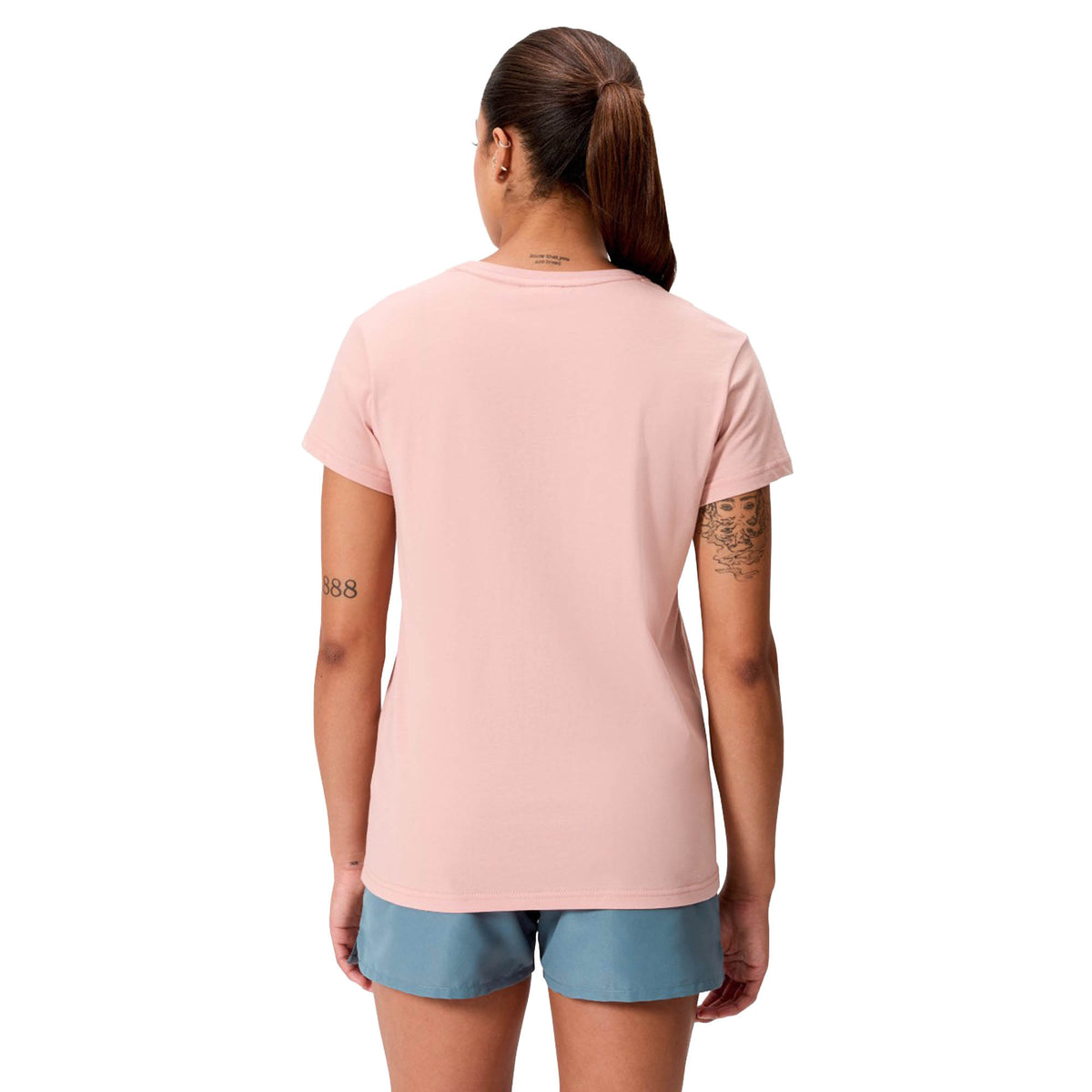 A person with long hair in a ponytail, wearing a pale pink Women’s Canterbury Uglies Logo T-Shirt by Canterbury and blue shorts, stands facing away from the camera against a plain background.