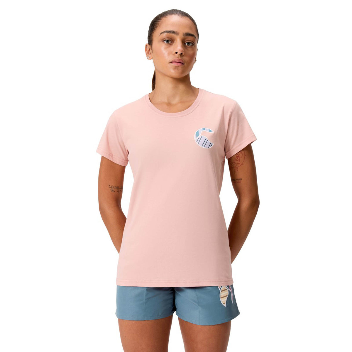 A person stands facing forward against a white background, wearing a light pink Canterbury Women’s Uglies Logo T-Shirt with a small chest graphic and blue shorts featuring a beige design.