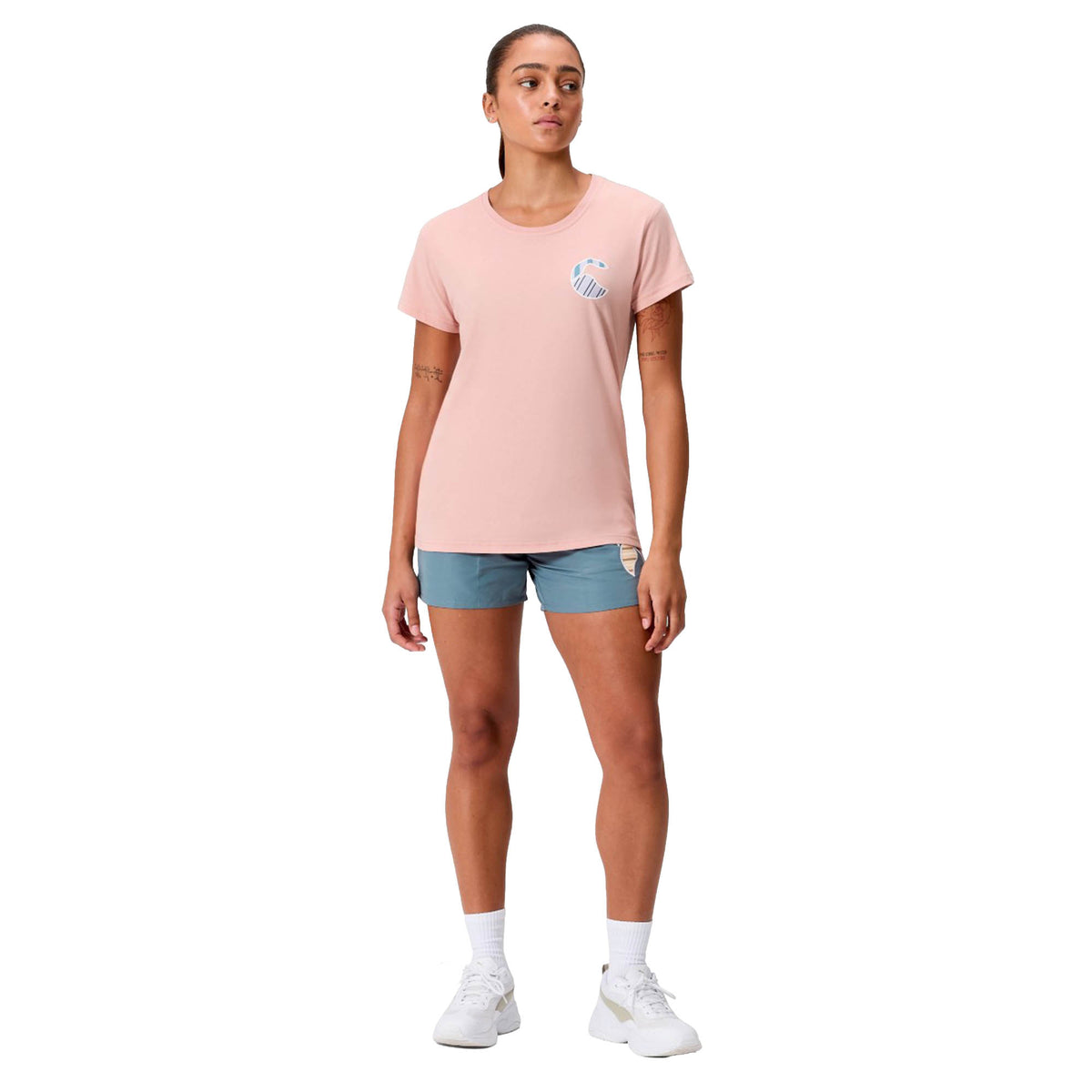 A person stands facing forward against a white background, wearing a pink Canterbury Women’s Uglies Logo T-Shirt, blue shorts, white socks, and white sneakers.