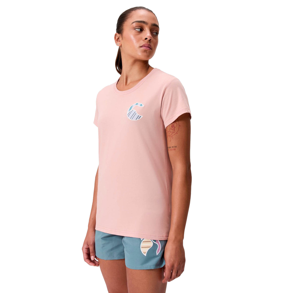 A person wearing a light pink Canterbury Women’s Canterbury Uglies Logo T-Shirt featuring a small chest graphic, paired with teal pastel-patterned shorts, stands against a white background.