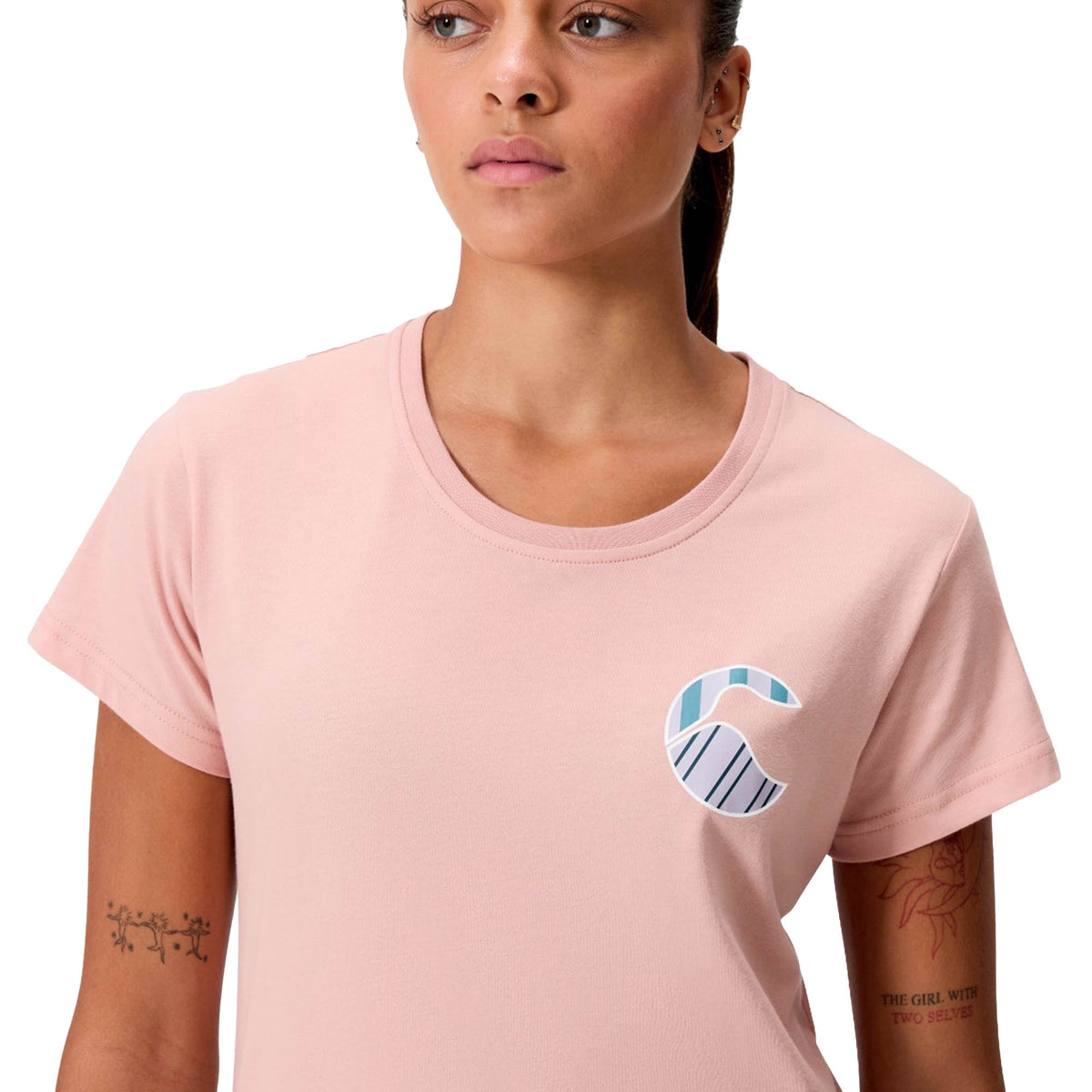 A woman wearing the Canterbury Women’s Uglies Logo T-Shirt, featuring a blue and white abstract “C” design on the chest, showcases visible arm and wrist tattoos against a white background.