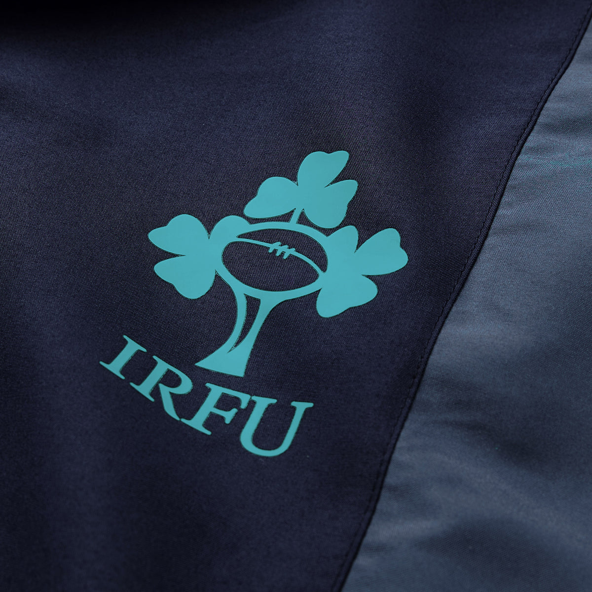 Close-up of a dark fabric showcasing the Ireland Rugby Football Union (IRFU) logo—three shamrocks and a rugby ball—printed in teal, as seen on the Canterbury Ireland 26 Legends 32″ Trackpant, an iconic piece for devoted Irish Rugby supporters.